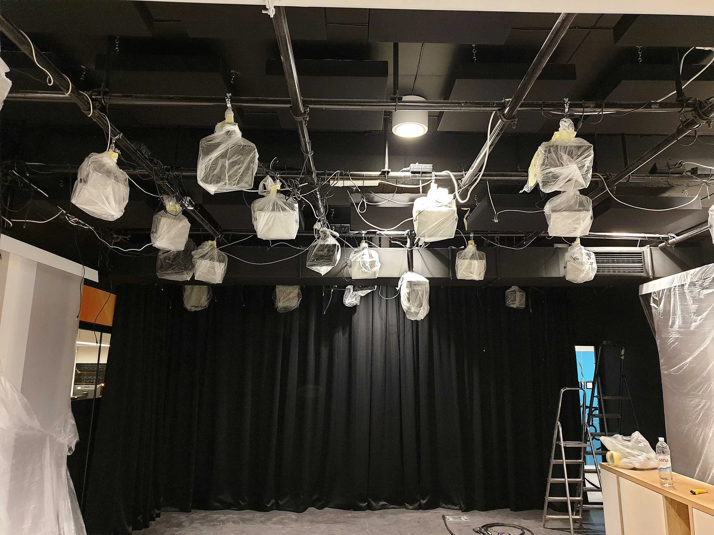Stage with black curtains in the background, ceiling lights, and hanging equipment covered in plastic bags. A ladder, a table with a water bottle, and other items are on the right.