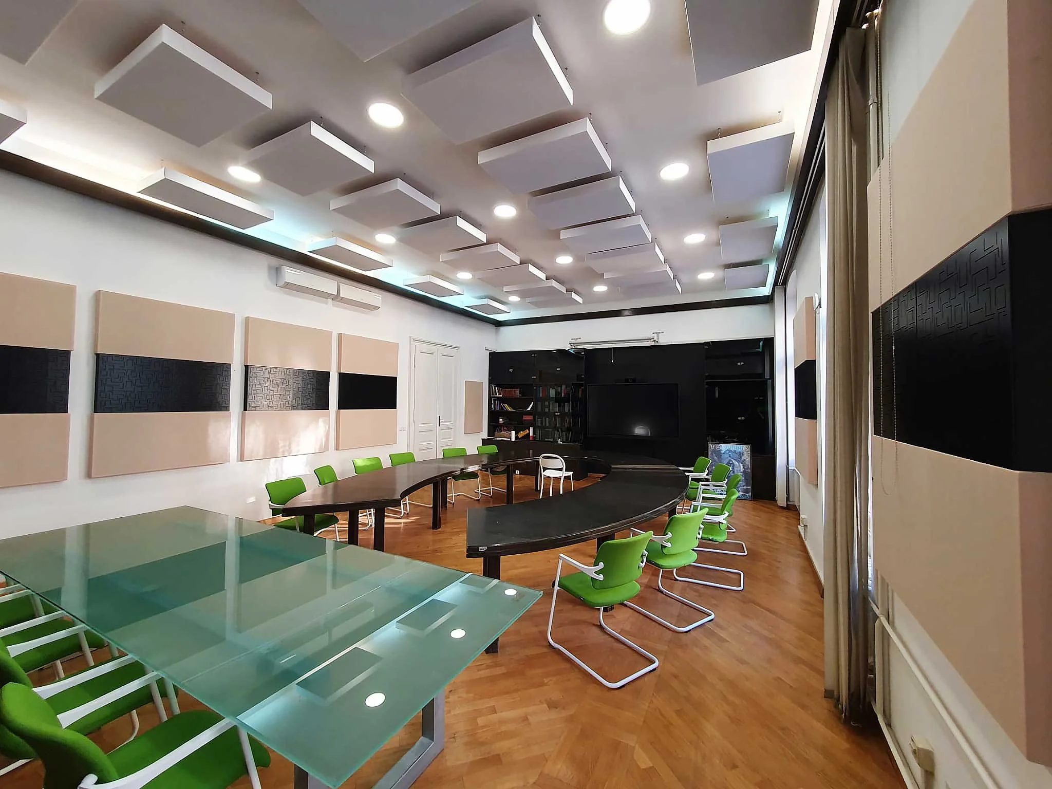 Empty conference room with green chairs, large curved table, soundproof panels on walls, and a black media wall with a TV in a modern office setting.