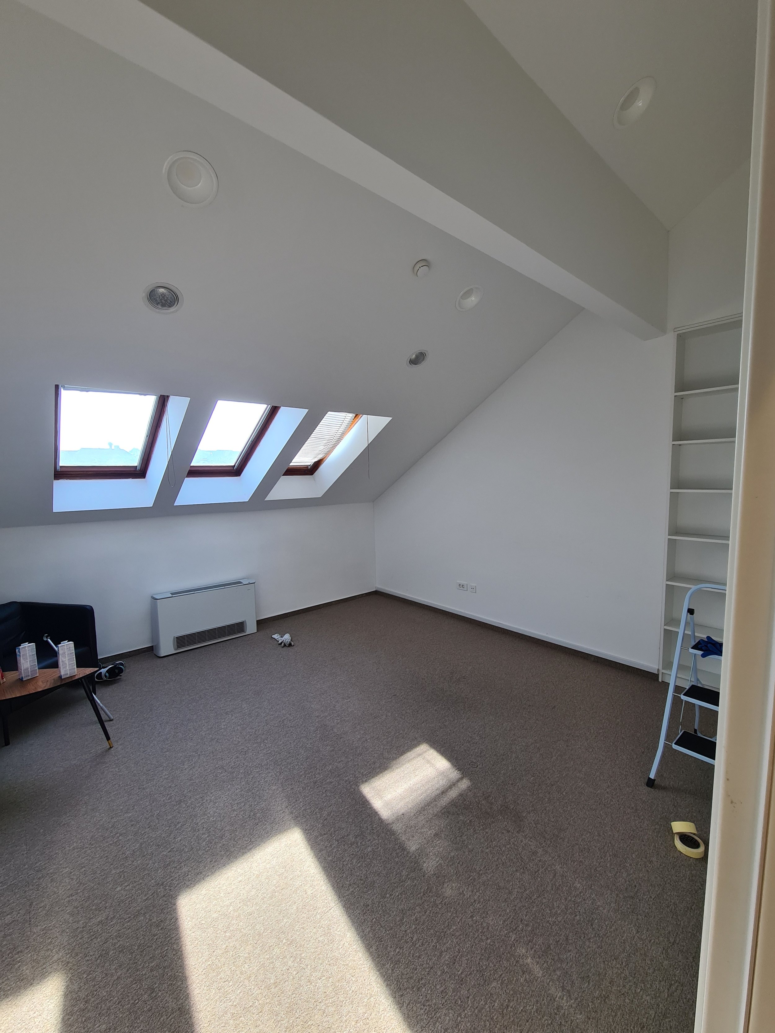 Empty attic room with beige carpet, three skylight windows on sloped ceiling, white walls, and a built-in shelf on the right. Sunlight streams in through the windows, casting a shadow on the floor. There are some scattered items including a small tab
