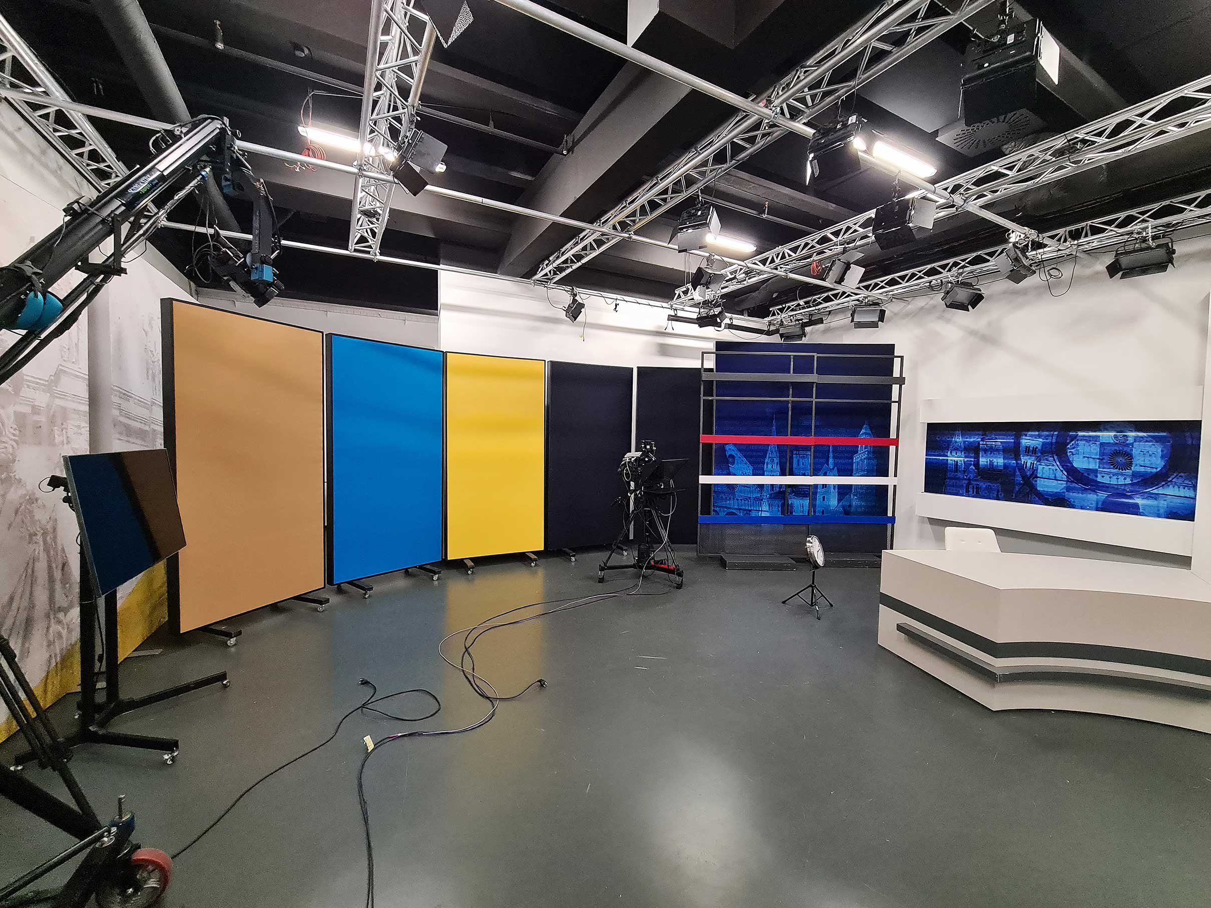 TV studio with multicolored partition walls, camera equipment, studio lights, and a news desk with a large digital screen.