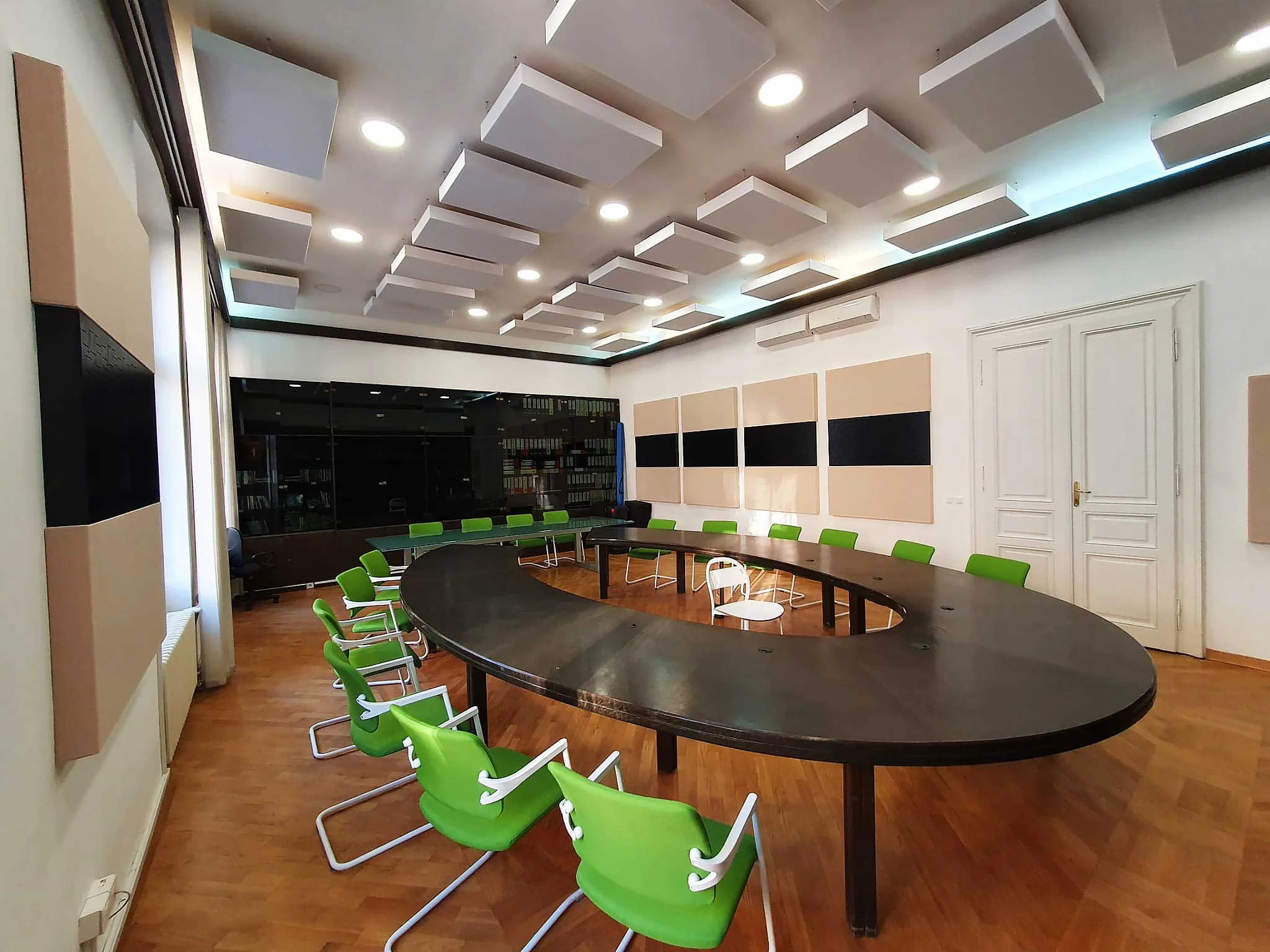 A modern conference room with a large, oval-shaped black table surrounded by bright green chairs. The ceiling has geometric white acoustic panels and recessed lighting. The room features soundproof wall panels and a white door.