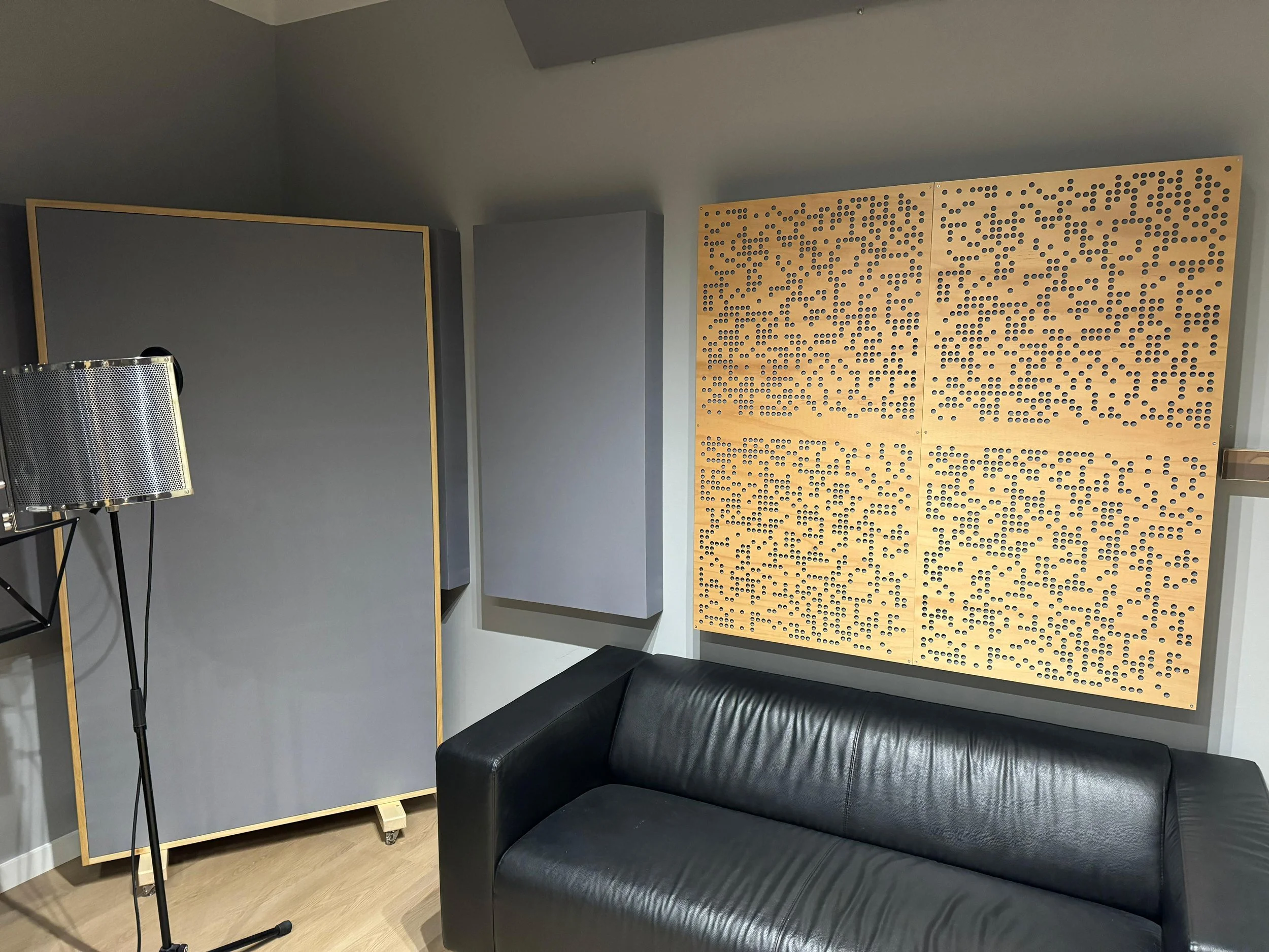 A modern music studio with black leather sofa, soundproof gray panels on the wall, and a large wooden acoustic panel with holes for sound absorption. There is also a standing microphone with a metal pop filter and a tall black stand.