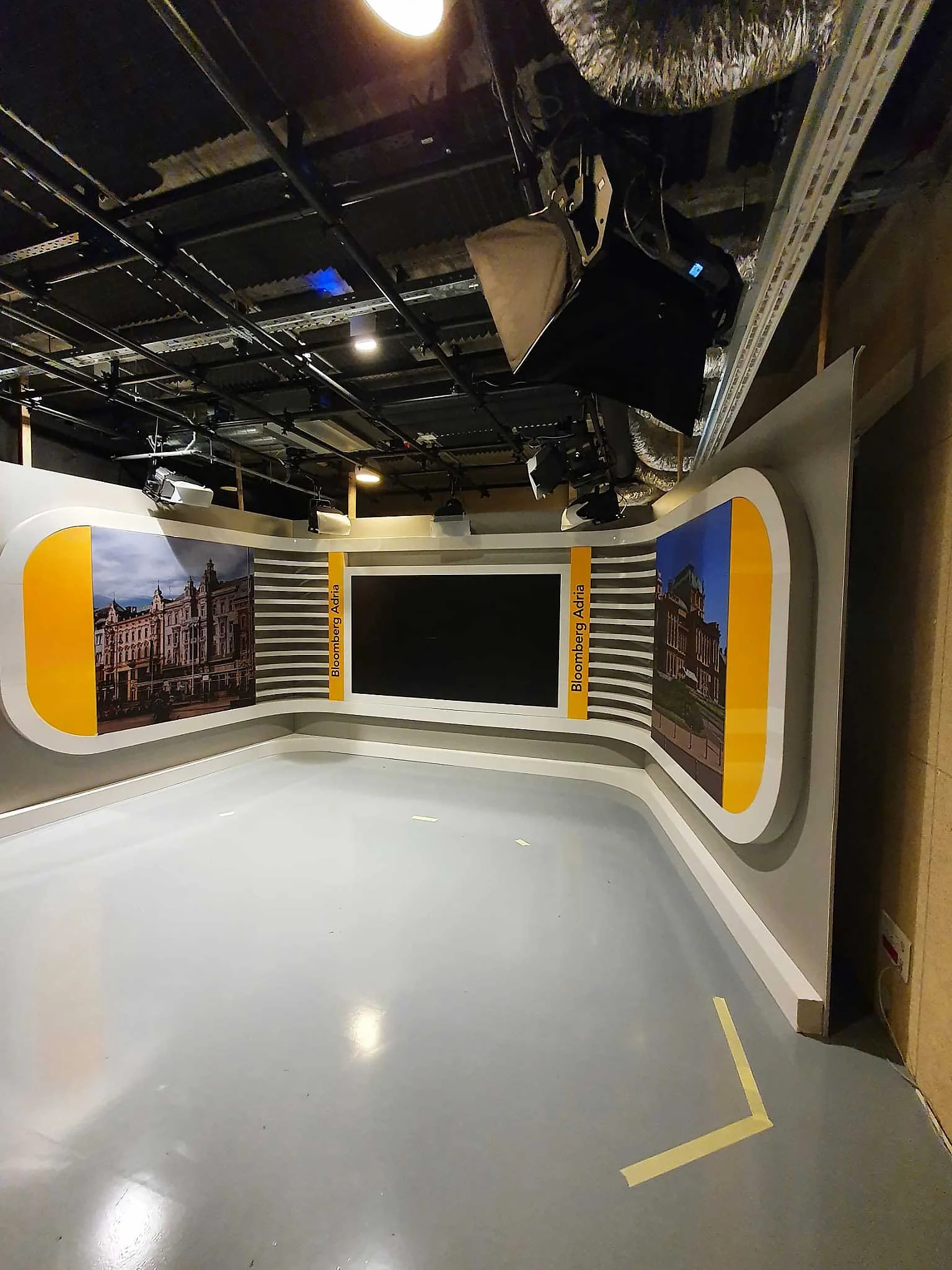 Empty television studio with a curved backdrop, two large screens showing historic buildings, and the text 'Bloomberg Adria' on yellow banners. Studio lights and equipment are overhead.