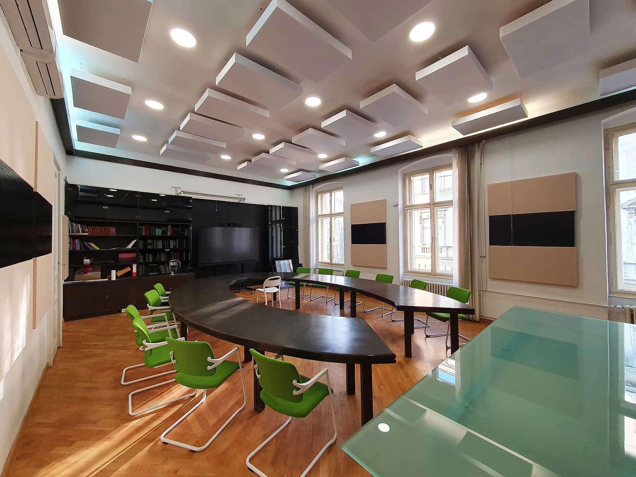 A conference room with a large, curved black table and several green chairs. The room has a bookshelf, a television, and large windows with light beige curtains. The ceiling has white rectangular acoustic panels and recessed lighting.