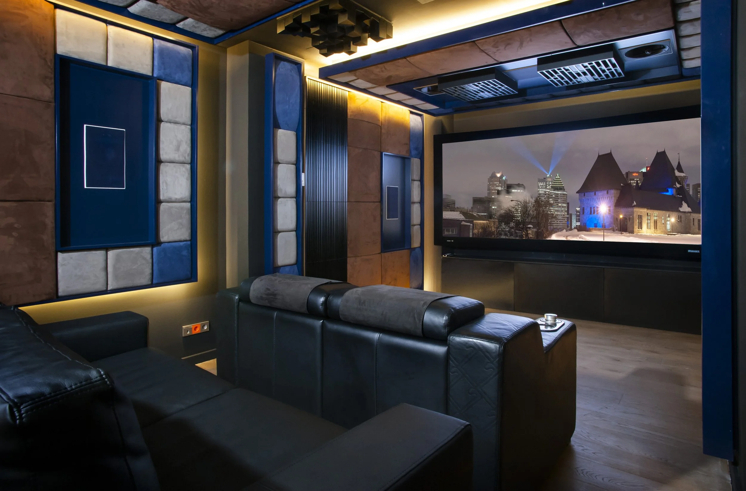 A modern home theater room with a large screen showing a cityscape with historic buildings at night, plush black leather seating, and soundproof wall panels with geometric patterns and soft lighting.