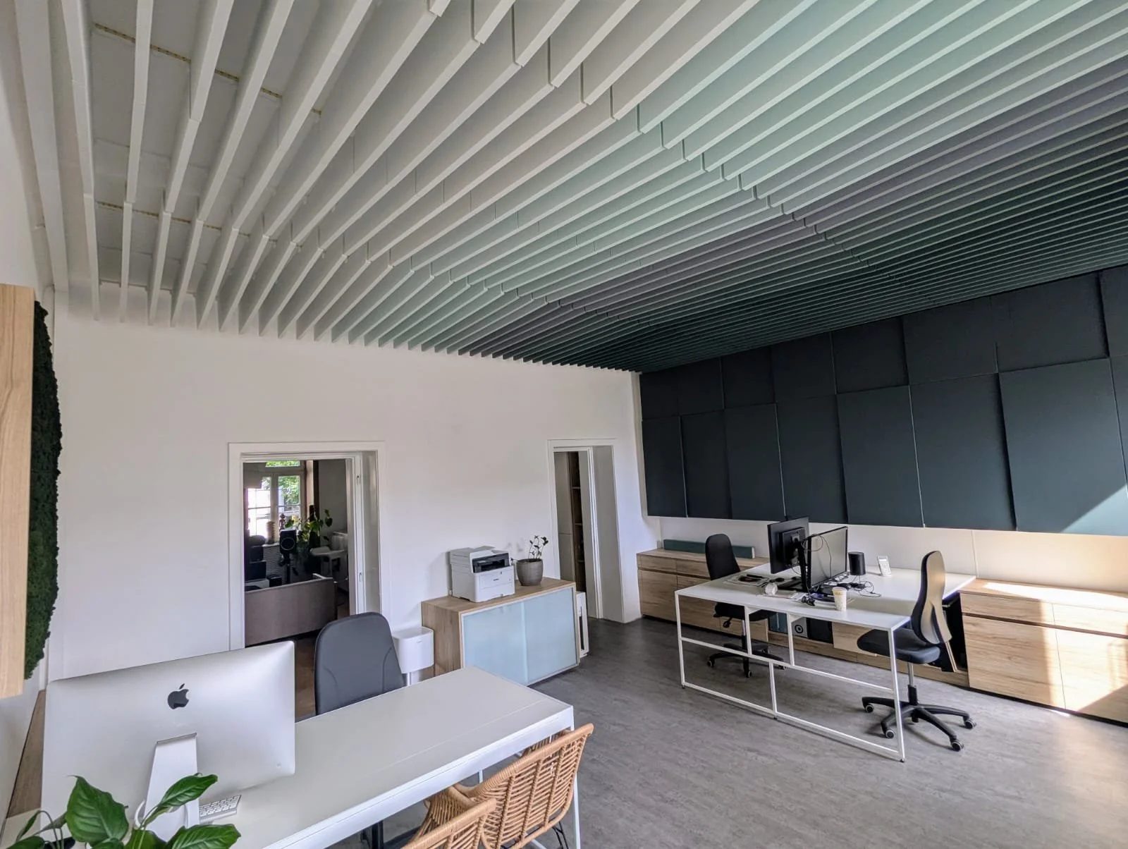 Modern office with white walls, wooden furniture, black wall panels, and a ceiling with white slats. There are desks with computers, a printer, and office chairs. A doorway shows another room with plants.