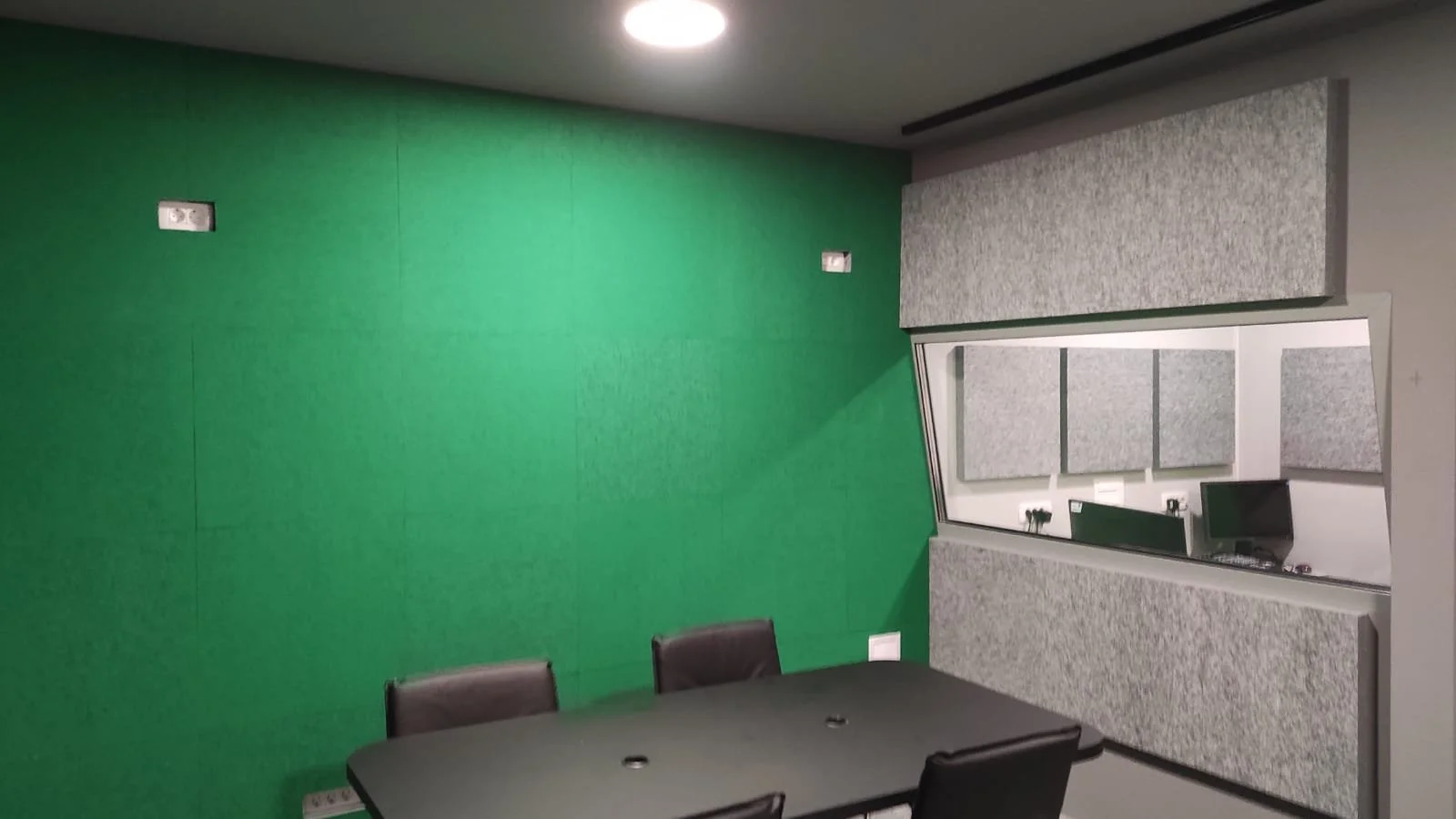 A modern conference room with a green accent wall, gray acoustic panels, a black conference table, and six black chairs, with a small window or mirror on the right wall and electronic equipment on a shelf.