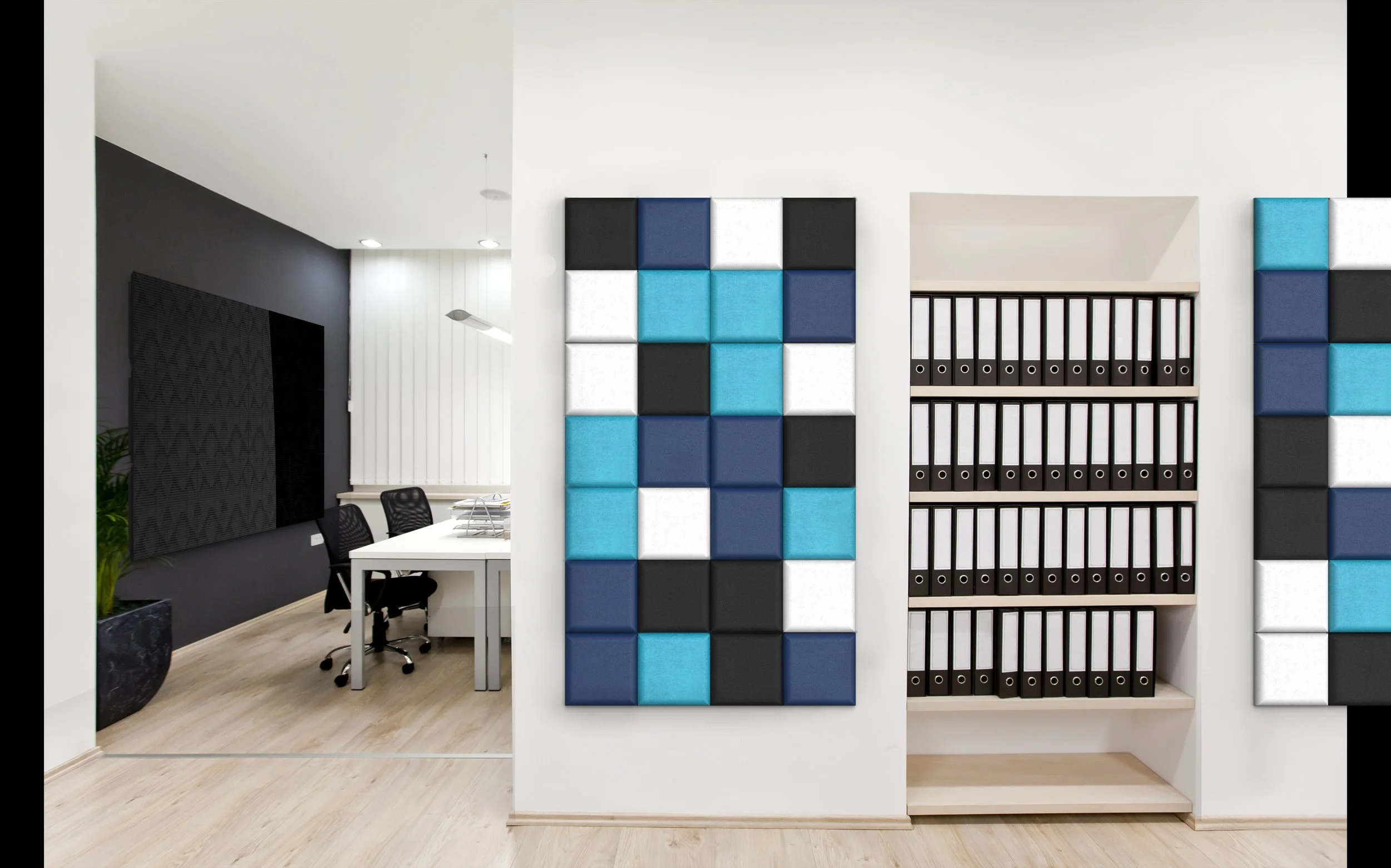 Office space with a white wall, decorated with blue, black, and white acoustic panels arranged in a grid pattern. To the right, there is a beige bookshelf filled with black ring binders. The background shows a meeting area with a white table, black c
