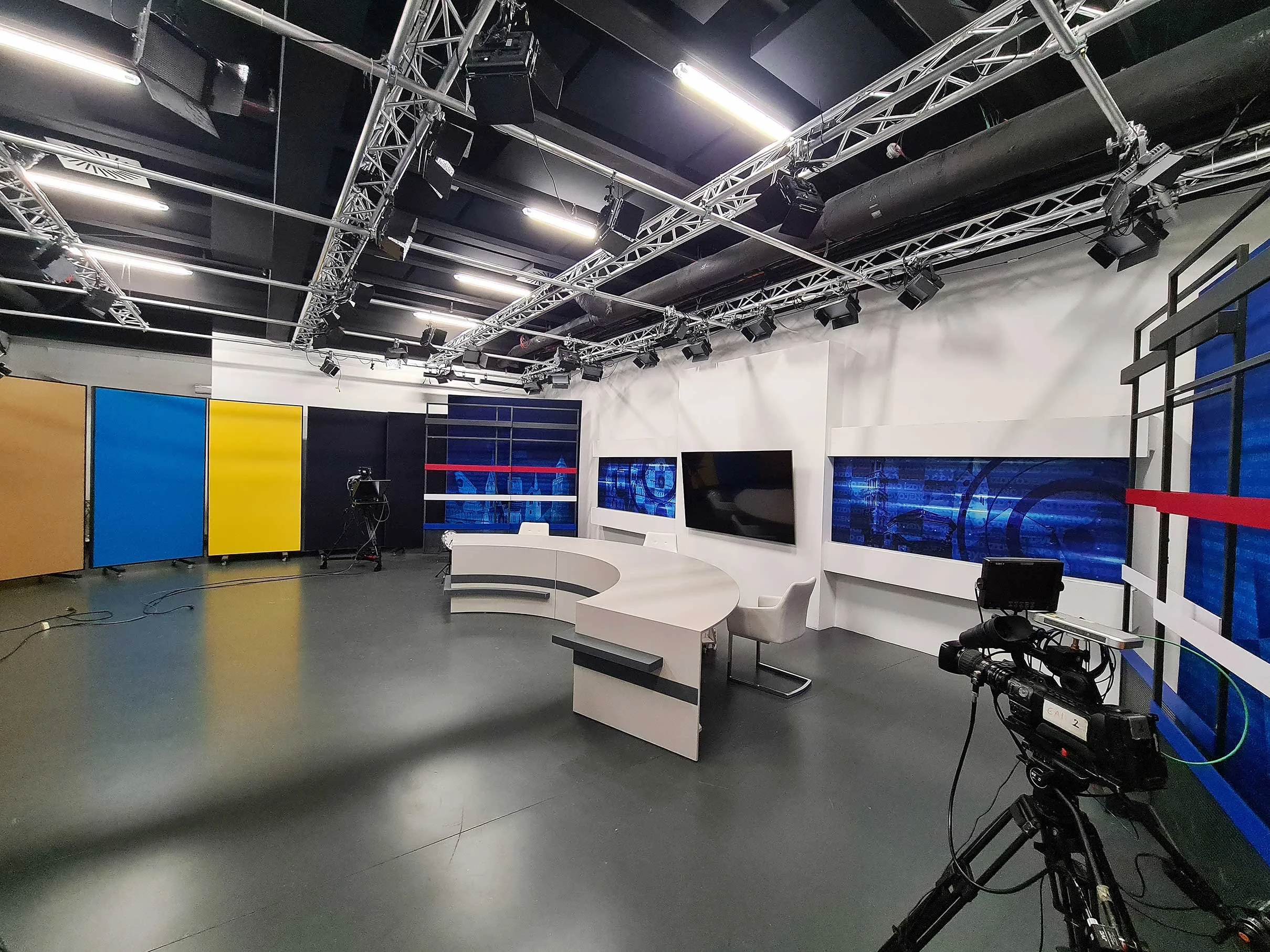 Empty television news studio with a curved desk, chairs, large monitors, and professional cameras.