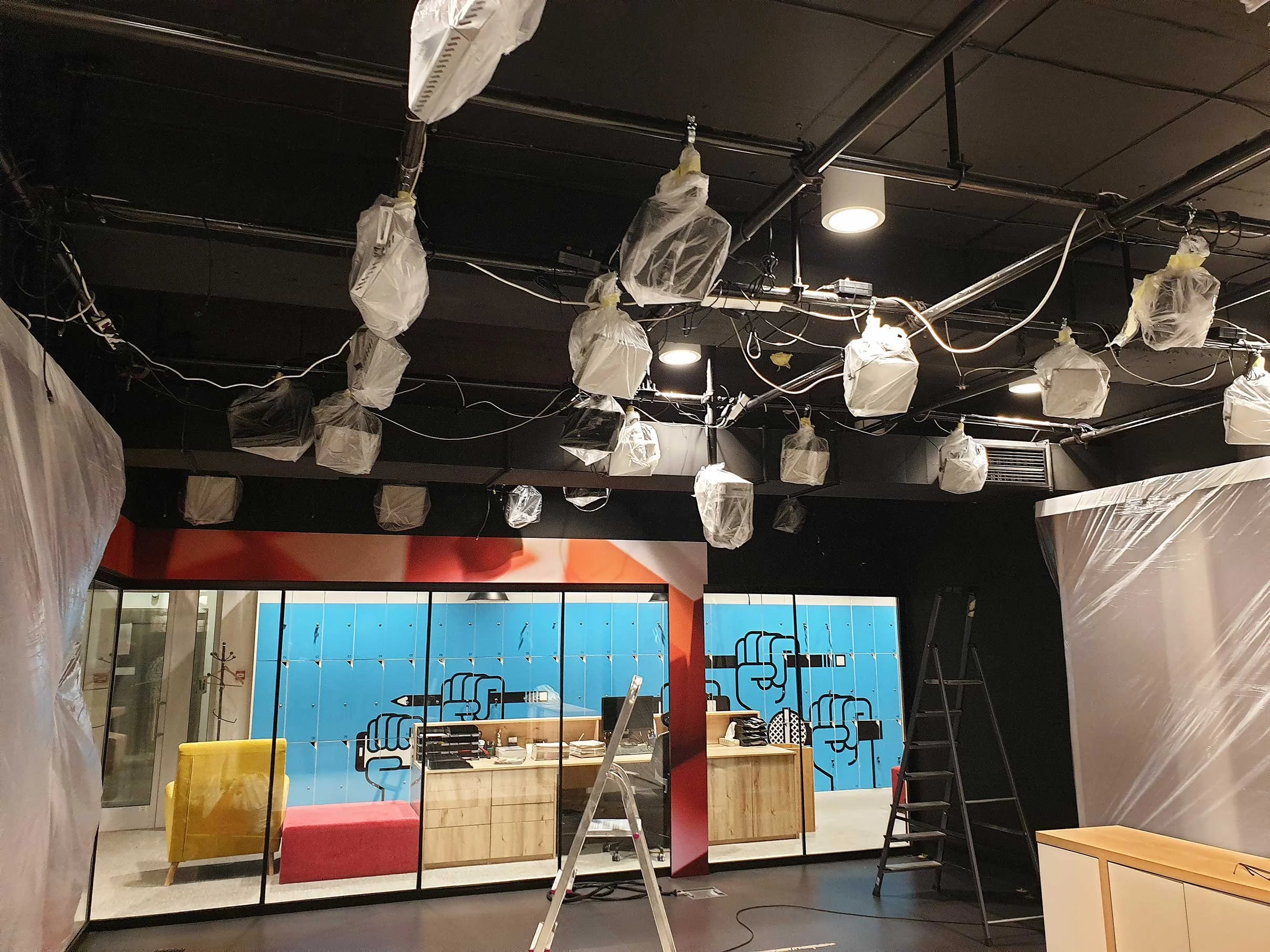 Interior of a space under construction with plastic-covered lighting fixtures on the ceiling, a ladder, a black wall, a glass partition, a colorful mural, and furniture including a yellow armchair and a red bench.
