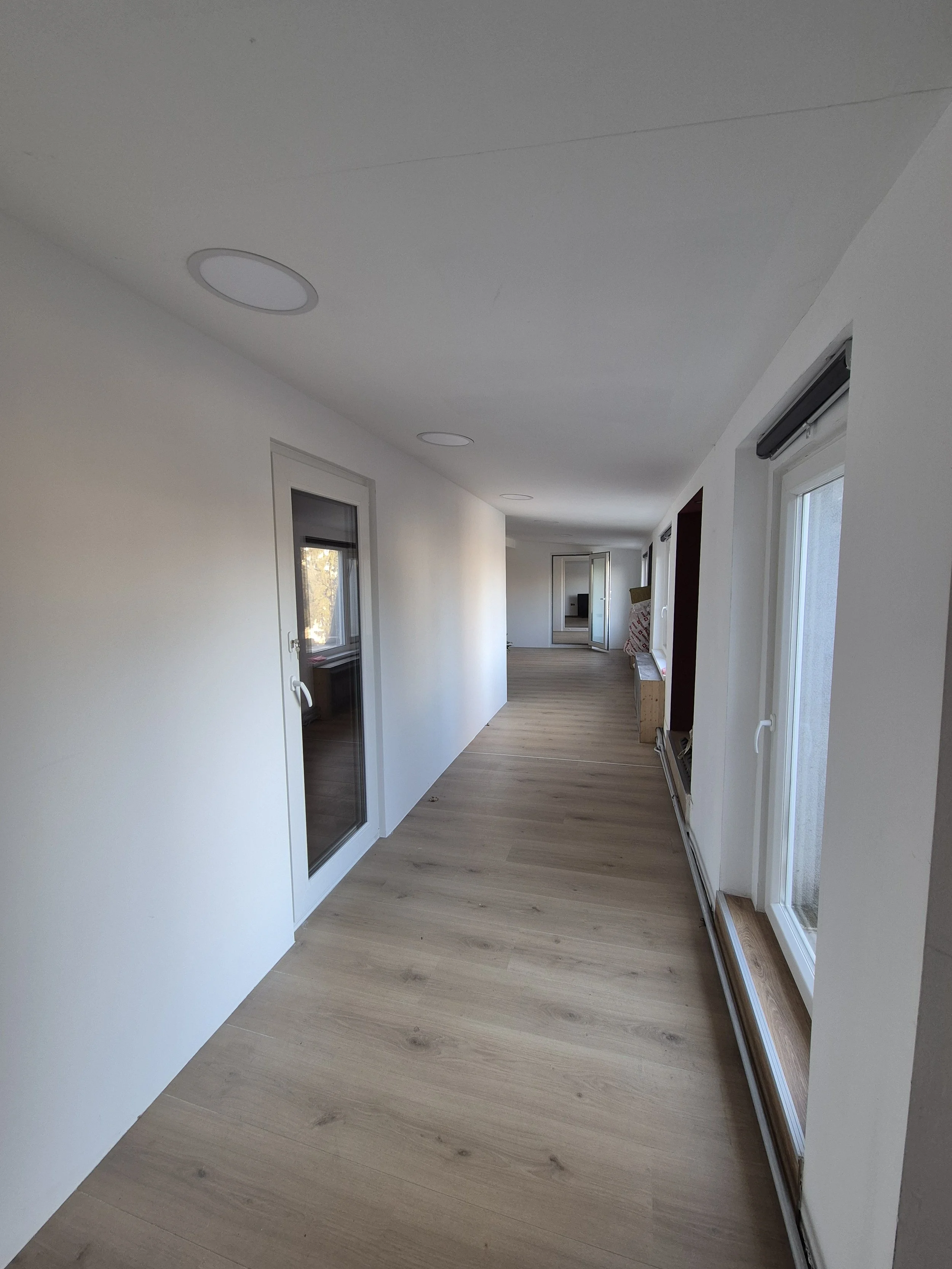 Empty hallway with white walls, wooden flooring, and large windows on the right side, some furniture and construction materials in the distance.
