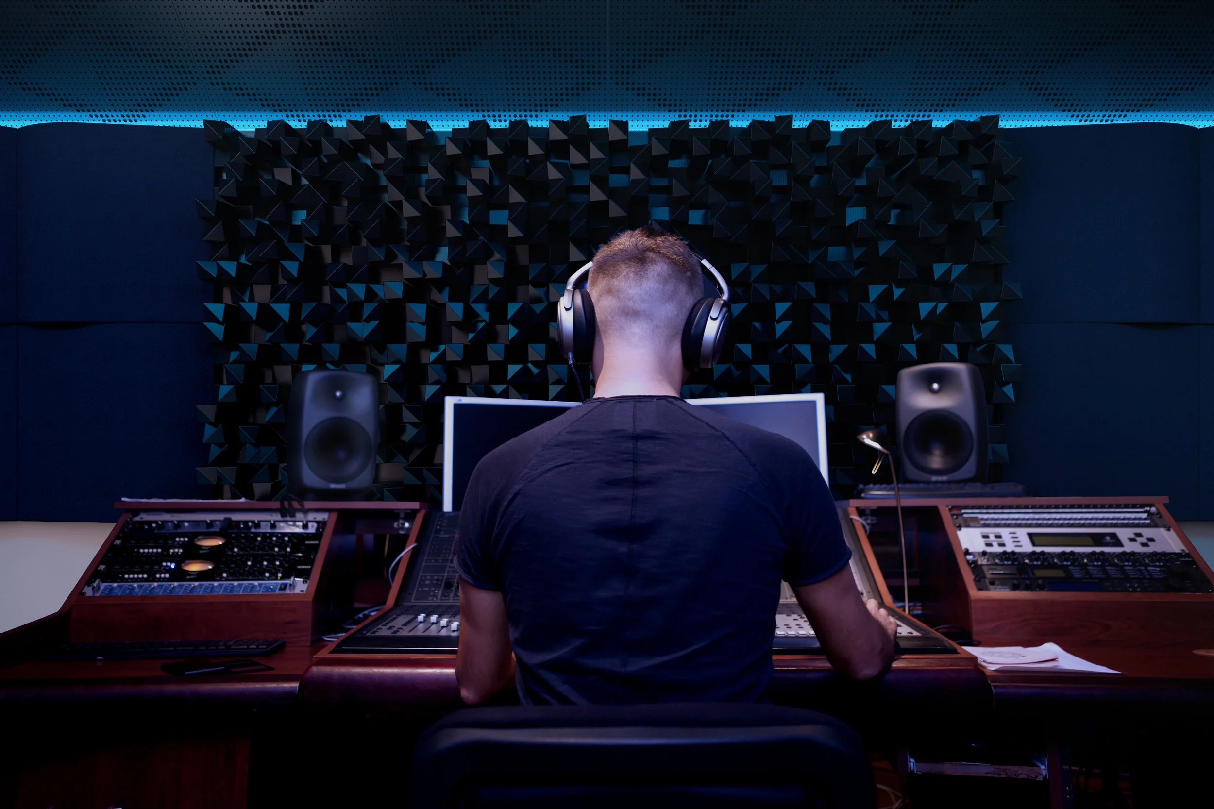 A person with short hair and a black T-shirt working in a recording studio, sitting at a mixing console with large studio monitors on either side, wearing headphones, with soundproof acoustic panels in the background.