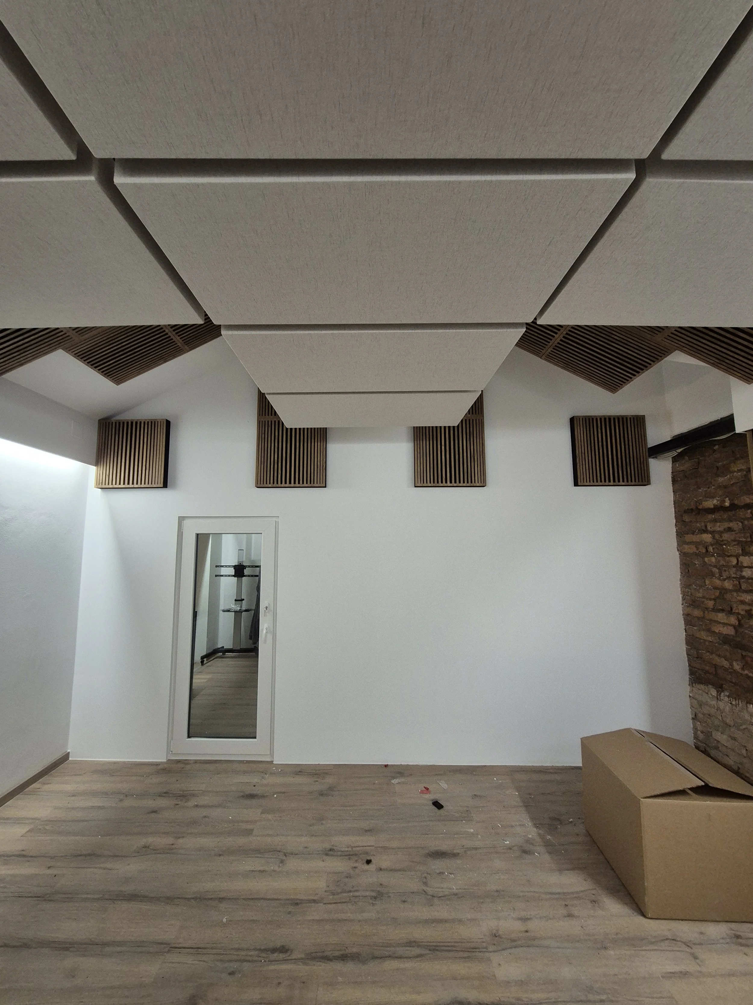 Empty room with wood floor, white walls, ceiling acoustic panels, and brick wall on the right. There is a mirror door and a cardboard box on the floor.