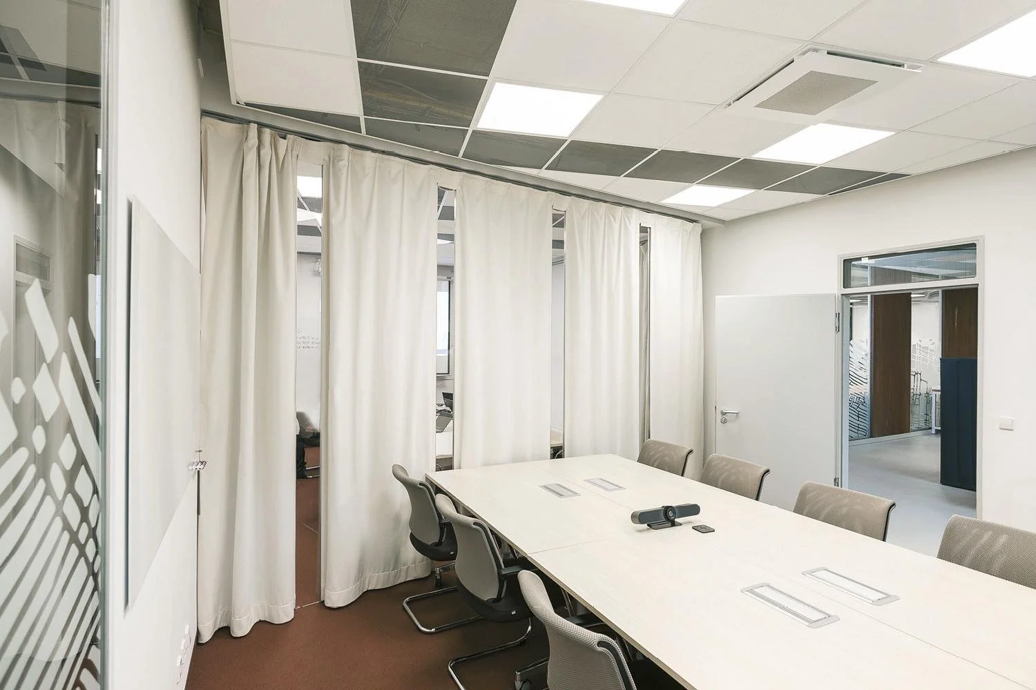 Empty conference room with white walls, a long white table, beige chairs, a camera on the table, and white curtains covering windows.
