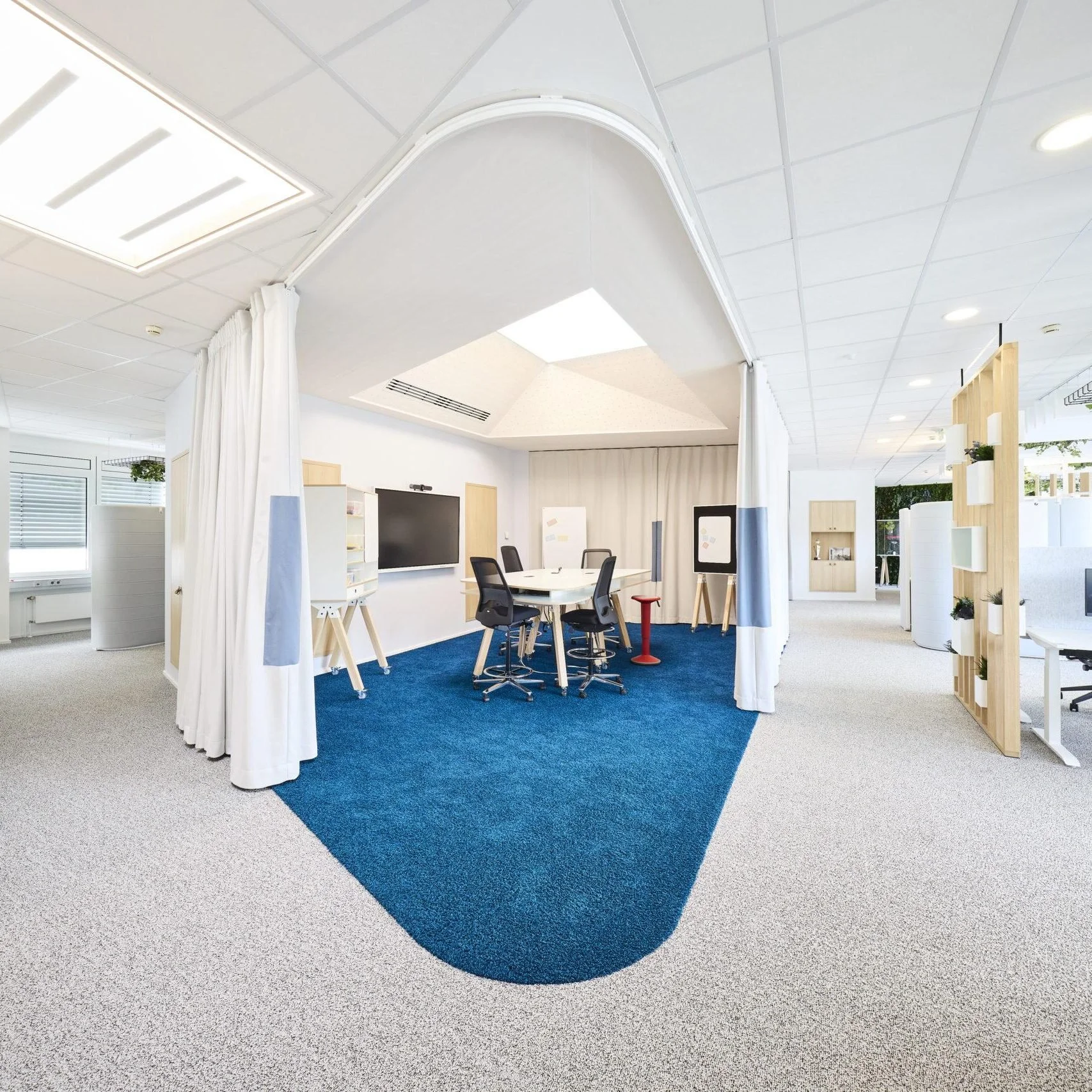 Modern office meeting room with a blue carpet, white walls, a round table surrounded by black chairs, a large black screen on the wall, and various whiteboards and shelves.