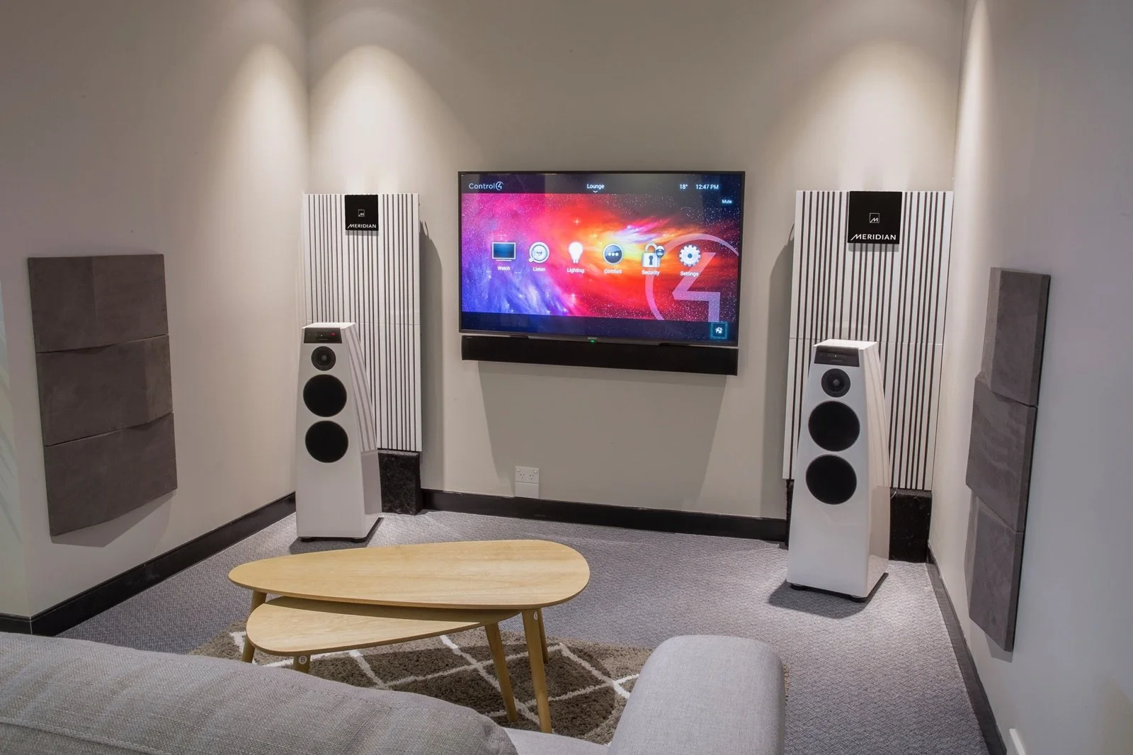 A modern lounge with a large flat-screen TV mounted on the wall, paired with two white floor-standing speakers and sound-absorbing panels on the walls. There is a small wooden coffee table and a gray upholstered sofa in the foreground.