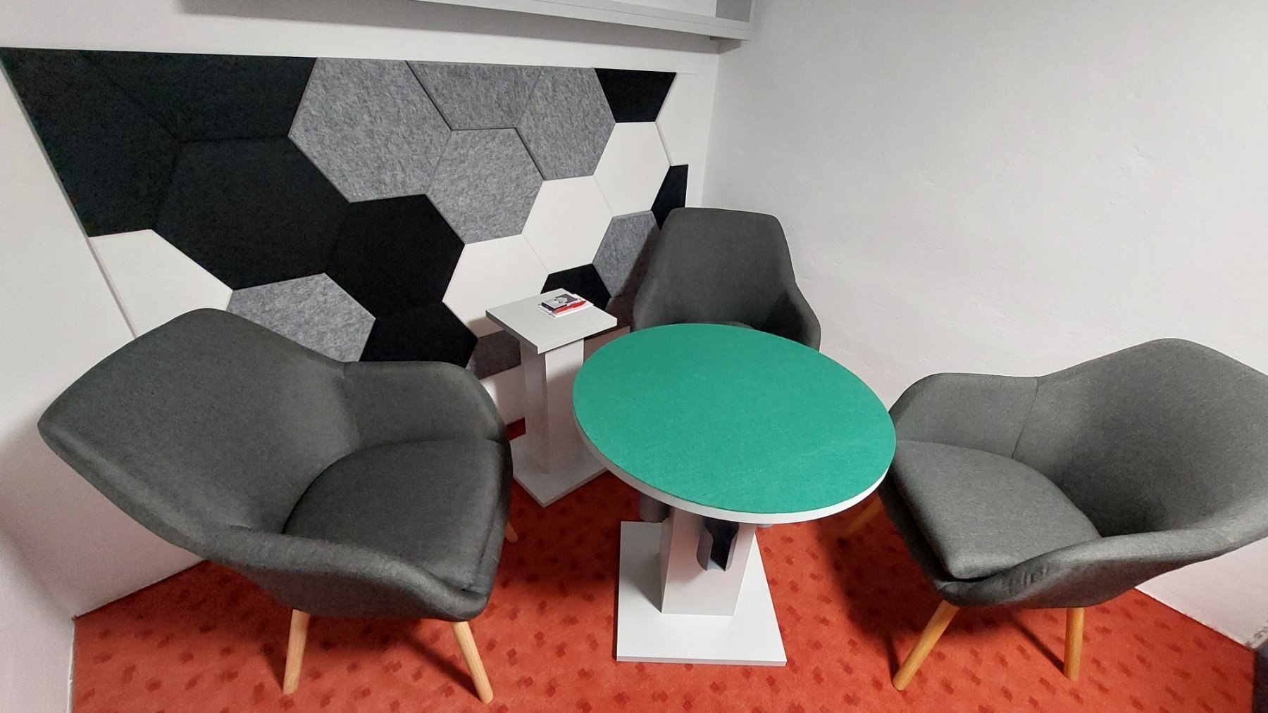 A small meeting area with three gray upholstered armchairs arranged around a small round table with a green top. There is a white side table with magazines between two of the chairs. The area has a decorative black, white, and gray hexagonal wall pan