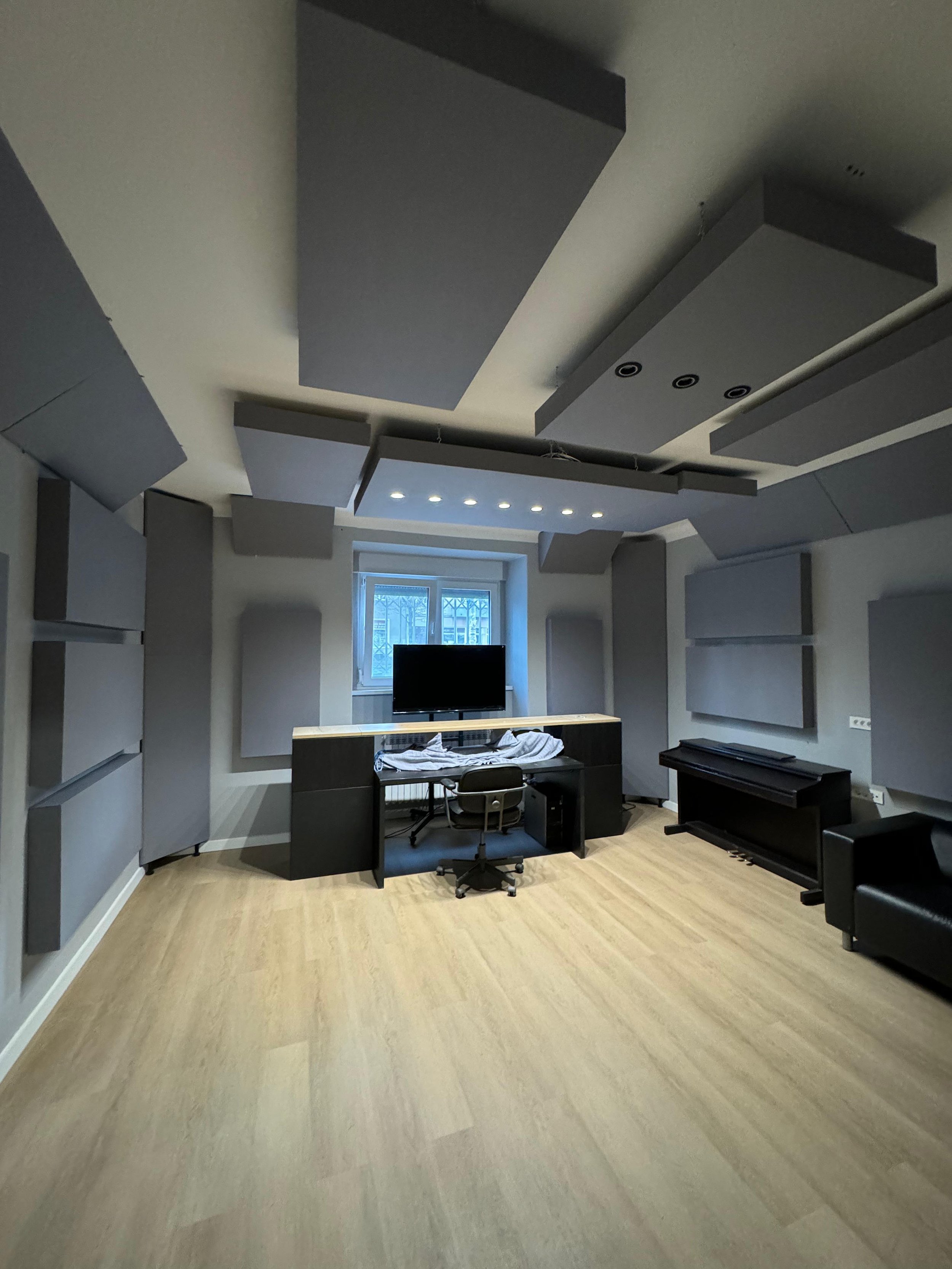 A recording studio with gray acoustic panels on the walls and ceiling, a window, a desk with a television, and a piano.