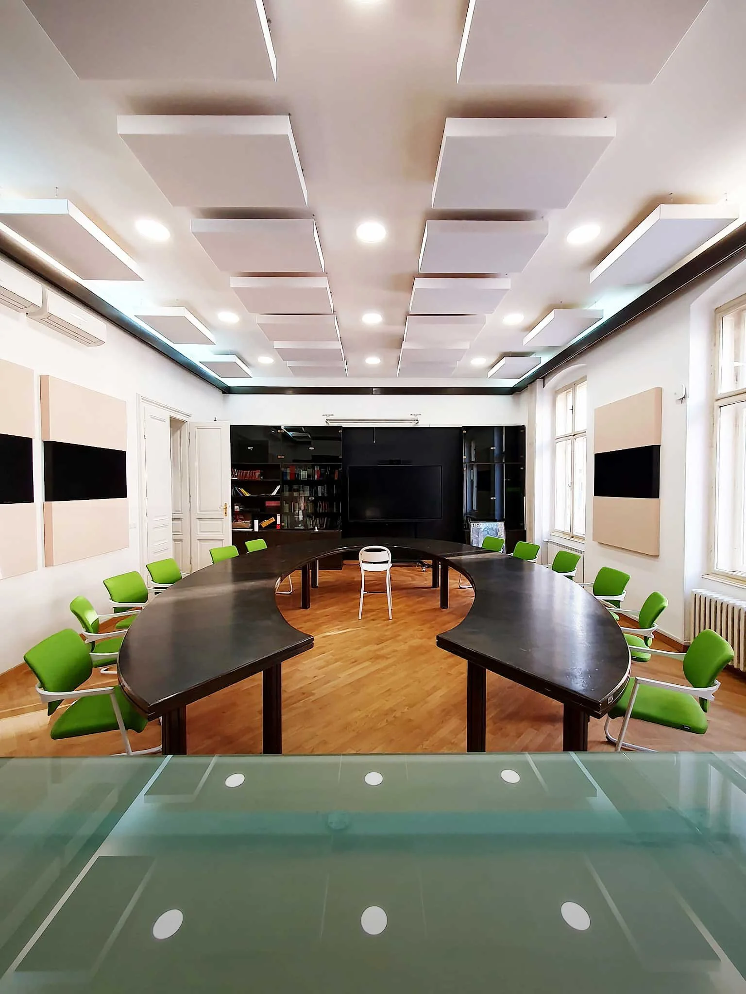 Empty conference room with a large U-shaped black table, green chairs, white walls, windows, and acoustic panels on the ceiling.