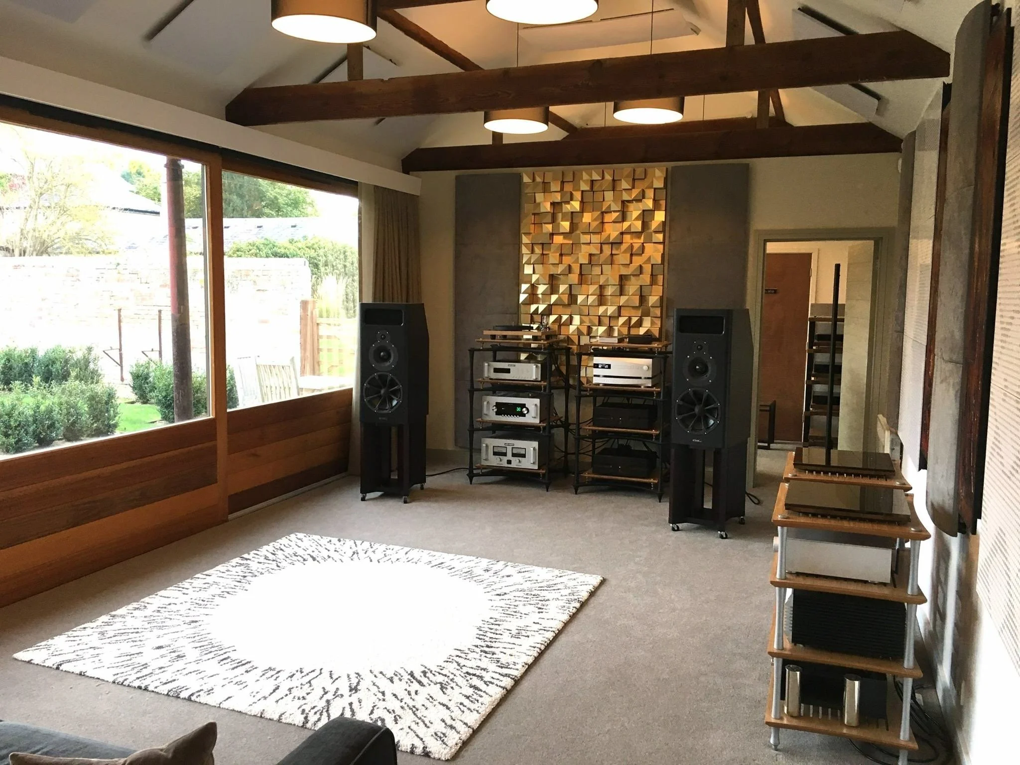A music listening room with large windows, beige carpet, a patterned area rug, two large black speakers, audio equipment shelves with silver components, acoustic panels on the wall, and a ceiling with exposed dark wooden beams.