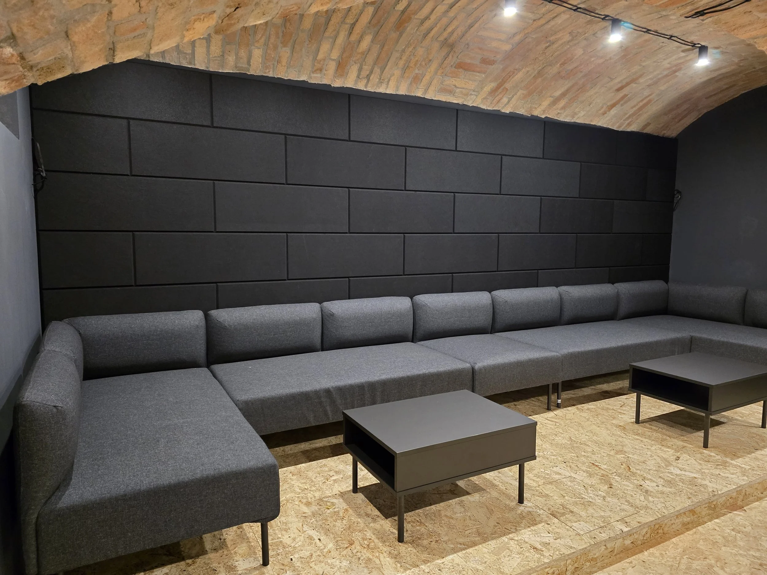 Modern lounge area with gray sectional sofa, two small black coffee tables, dark wall panel, and brick arched ceiling with track lighting.