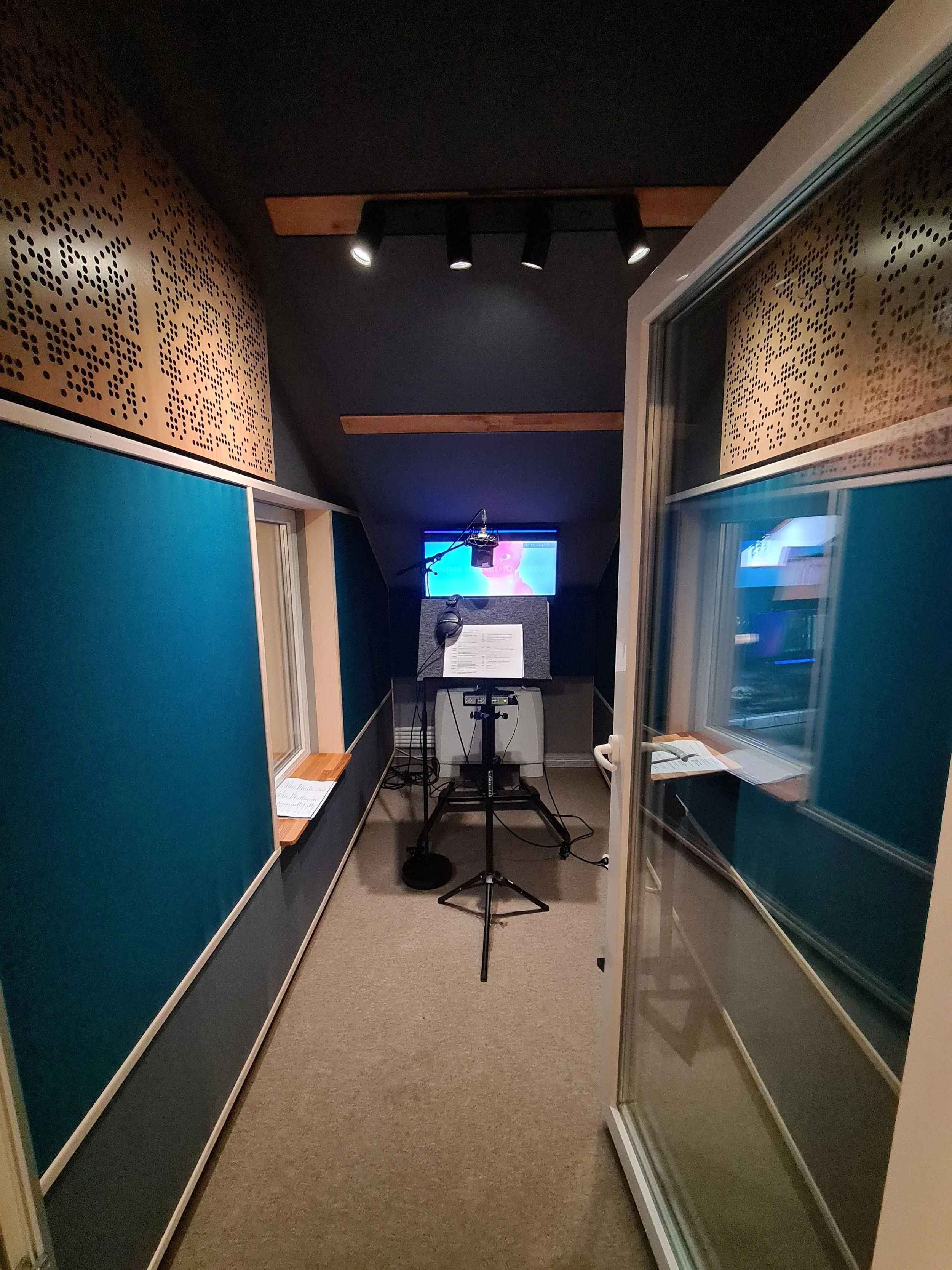 Recording studio vocal booth with microphone, music stand, headphones, and a monitor with a music video playing.