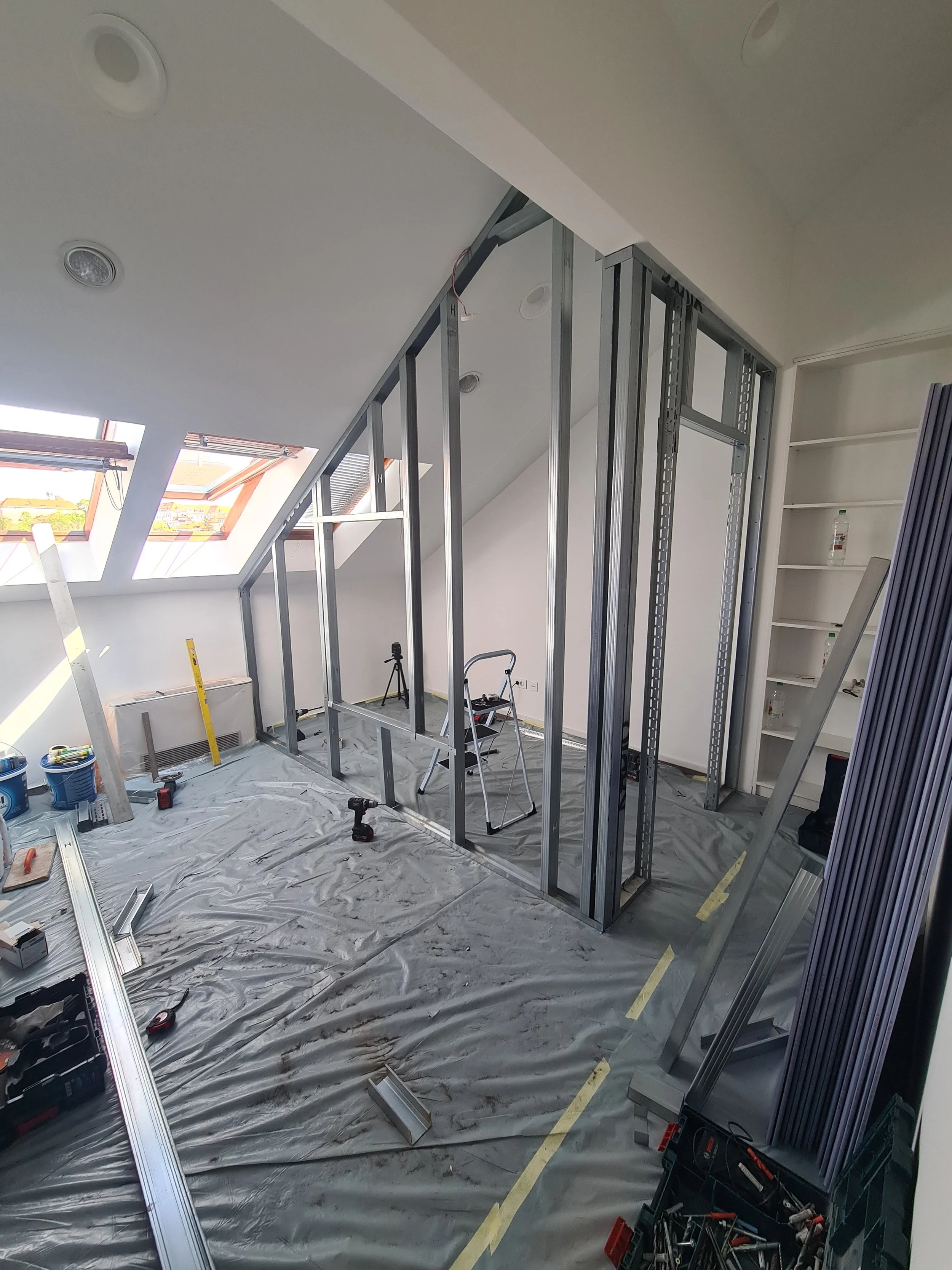 Construction site with metal wall framing inside a room with skylight windows on the sloped ceiling, construction tools and materials on the plastic-covered floor.