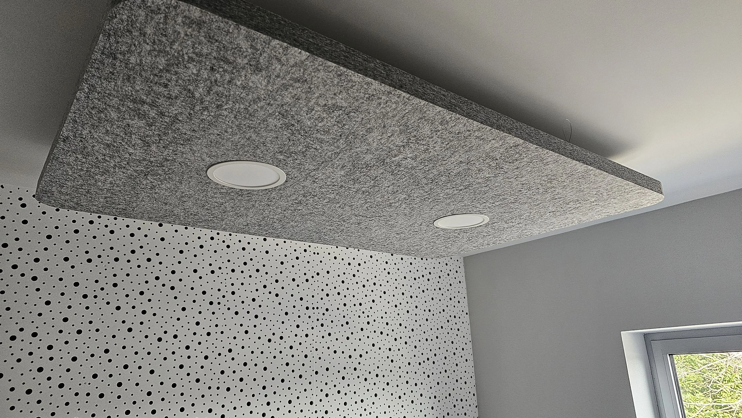 Ceiling with grey acoustic panels, two installed round recessed lights, corner of a room with a white wall and a window showing trees outside.