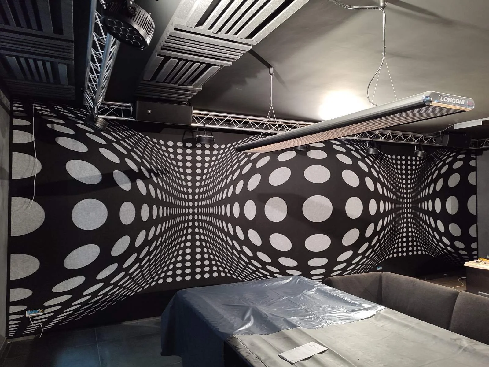 Room with a black wall featuring a optical illusion design of swirling white circles, a table covered with a plastic sheet, and a black couch. There is a Longoni light fixture hanging from the ceiling, with various electronic and lighting equipment m