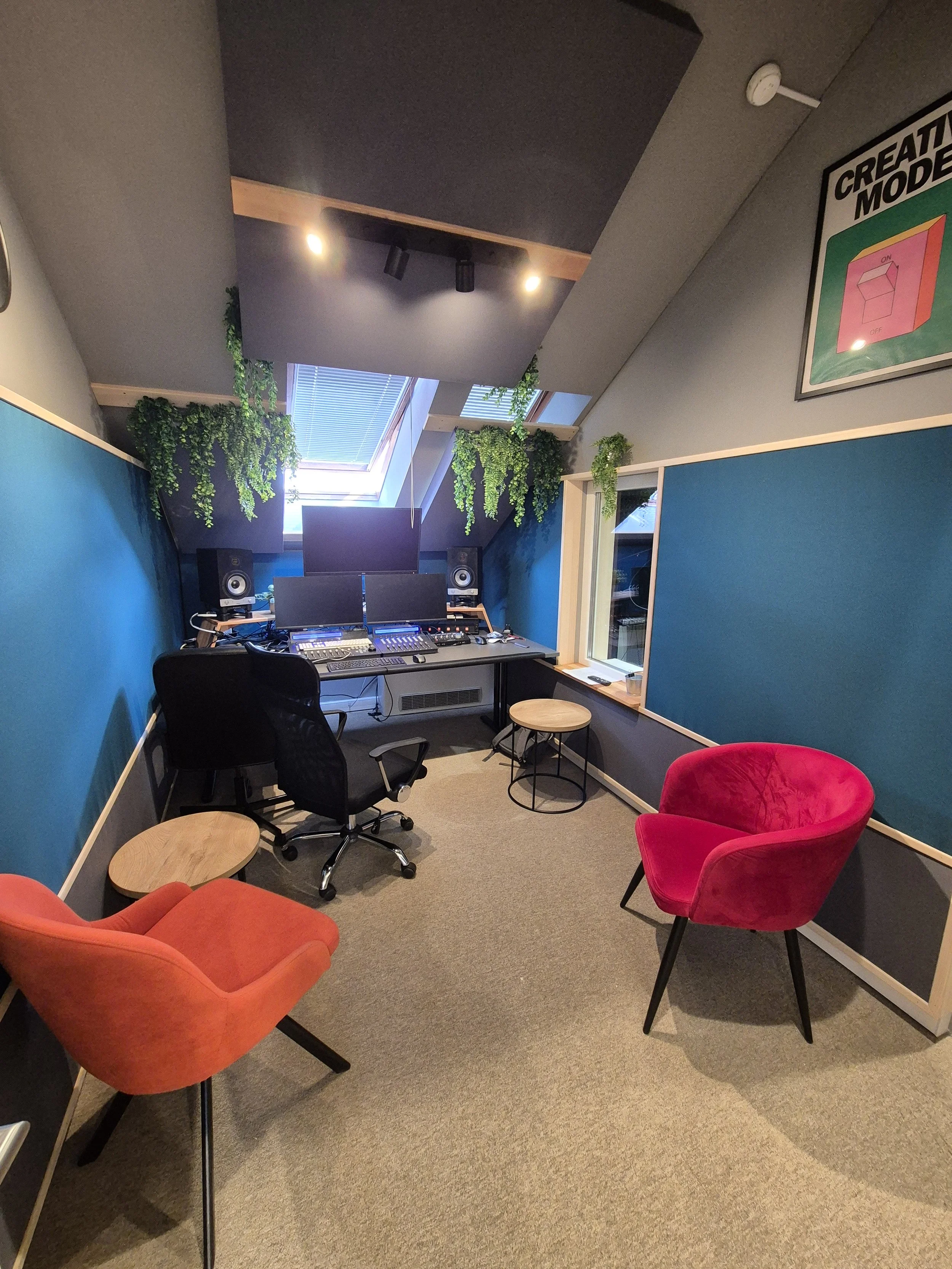 A small music or recording studio with a desk featuring multiple monitors, studio speakers, and audio equipment. There are colorful chairs, potted plants hanging from the ceiling, a skylight, and a framed poster on the wall.