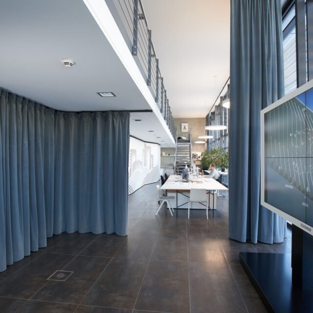 Modern office space with a staircase, large windows, blue curtains, and a meeting table with chairs.