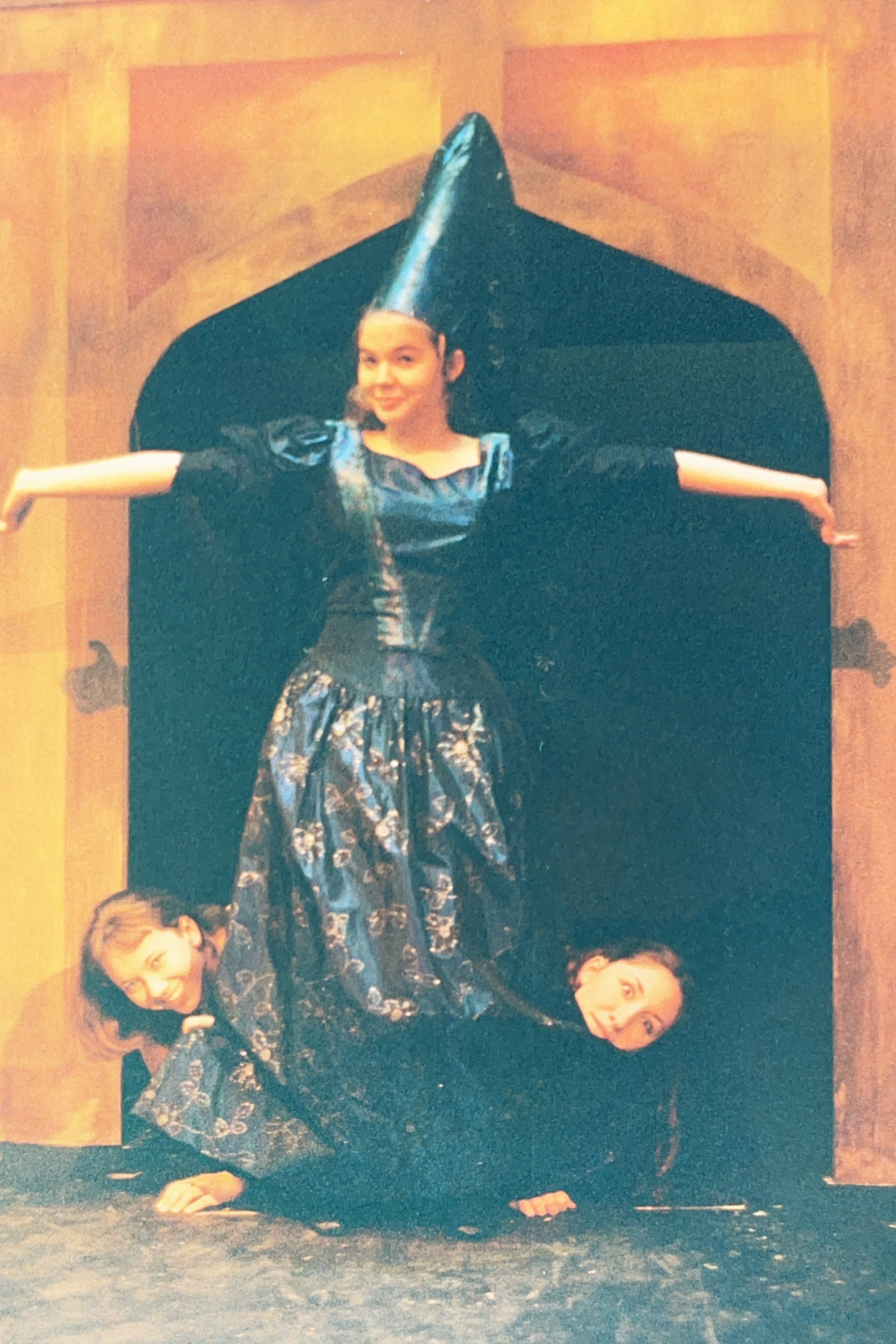 Three people in theatrical costumes performing on stage. The central figure wears a tall black hat and a long blue gown. Two others are crouched below, peeking out from under the gown.