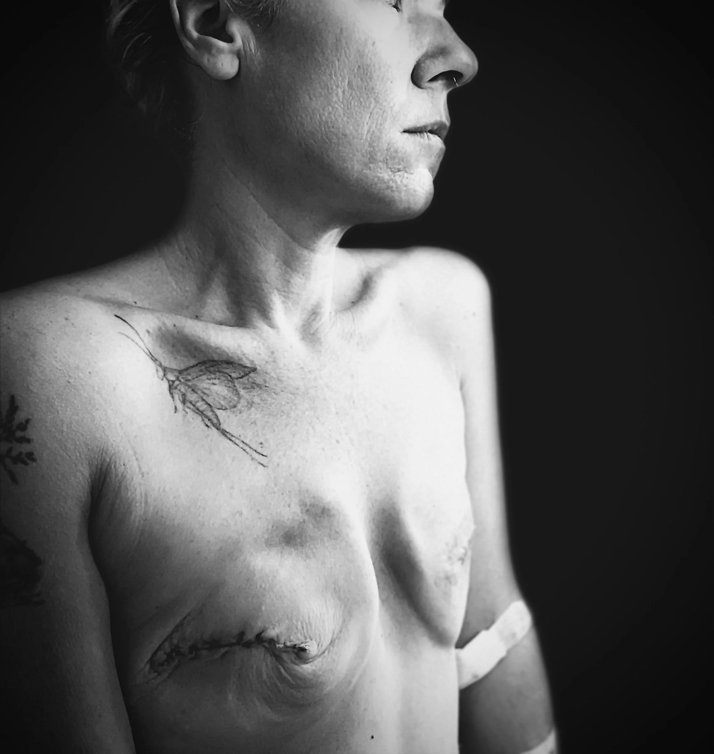 Black and white photo of a topless woman with tattoos on her shoulder and chest, looking to the side with a serious expression.