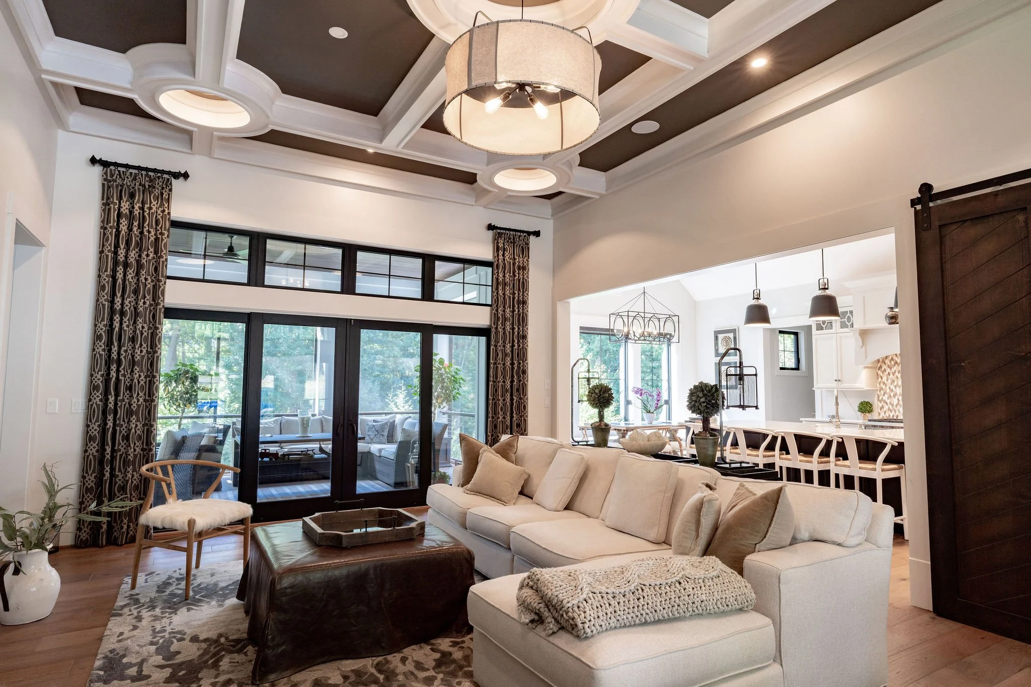 Living room with a white sectional sofa, decorative pillows, and a large ottoman. Behind the sofa, a bar counter with bar stools is visible. In the background, sliding glass doors lead to an outdoor patio with seating, and a dining area with a chandelier. The ceiling features coffered details and a modern drum light fixture. Large windows and drapes add natural light to the space.