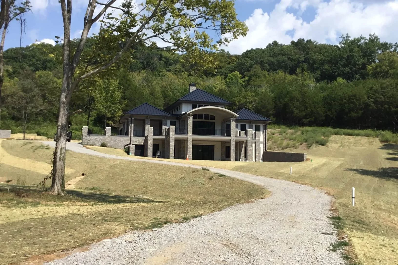 Modern new construction two-story house with dark roof, large windows, and a gravel driveway on a grassy hill with trees behind.