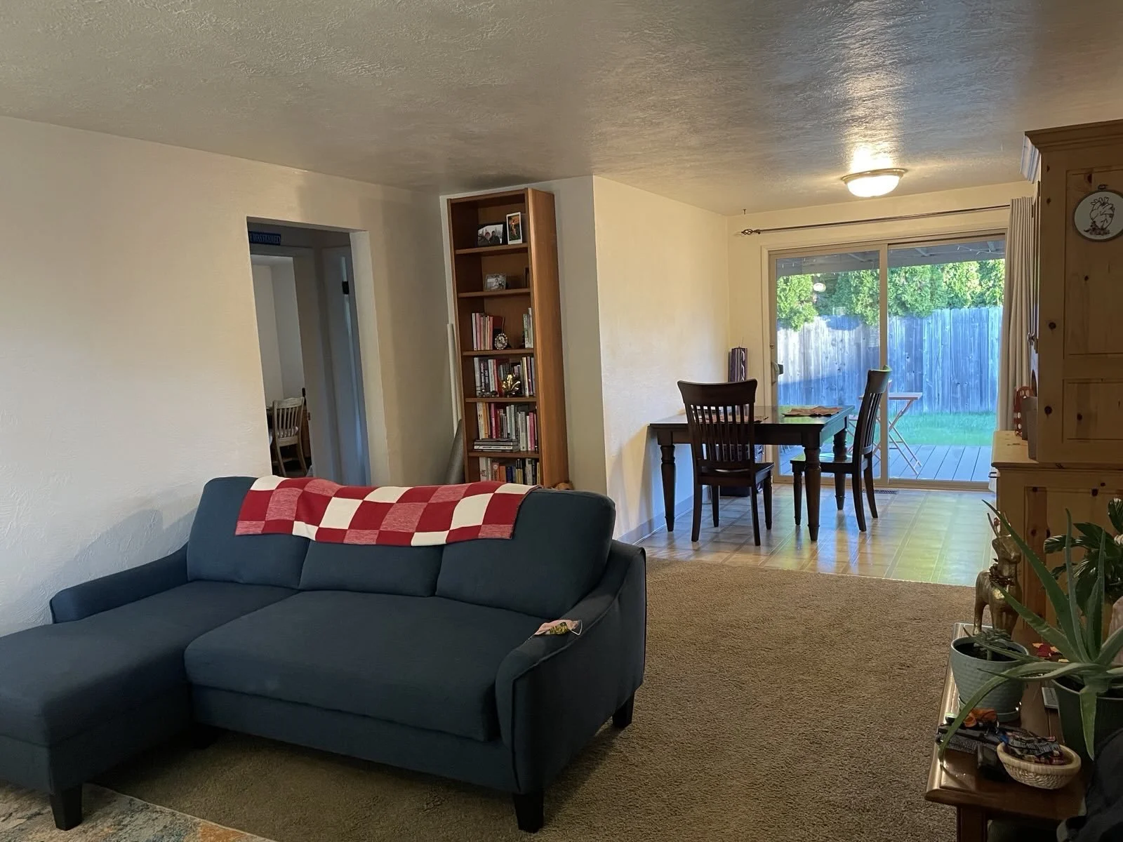 Living room with a blue sectional sofa with a red and white checkered blanket, a brown bookshelf filled with books and framed photos, a dining table with chairs near a sliding glass door leading to an outdoor patio, and a houseplant on a wooden side 