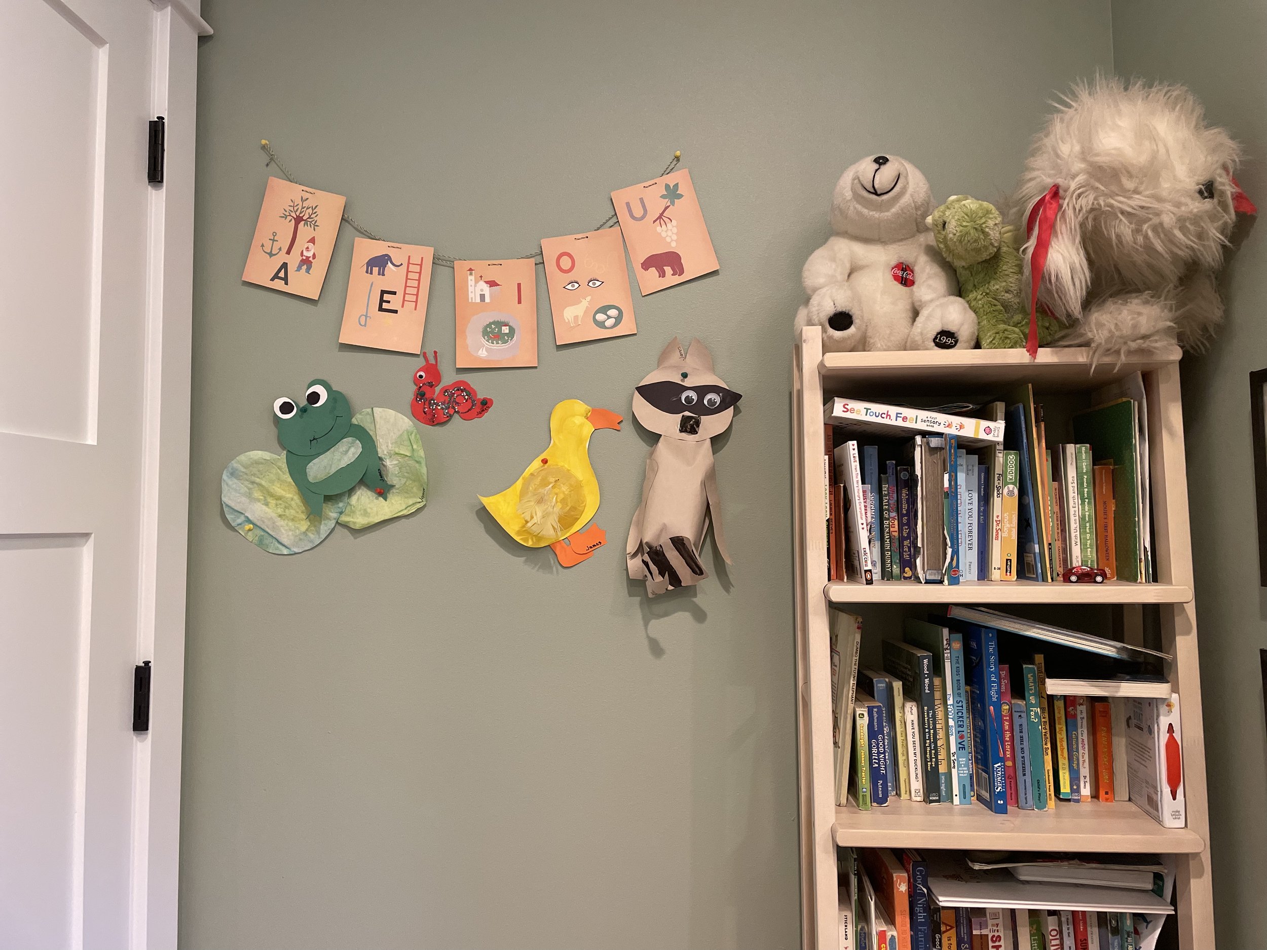 Decorative wall with a banner spelling 'ALOHA' and various paper and plush animal crafts, a bookshelf with books and stuffed animals on top.