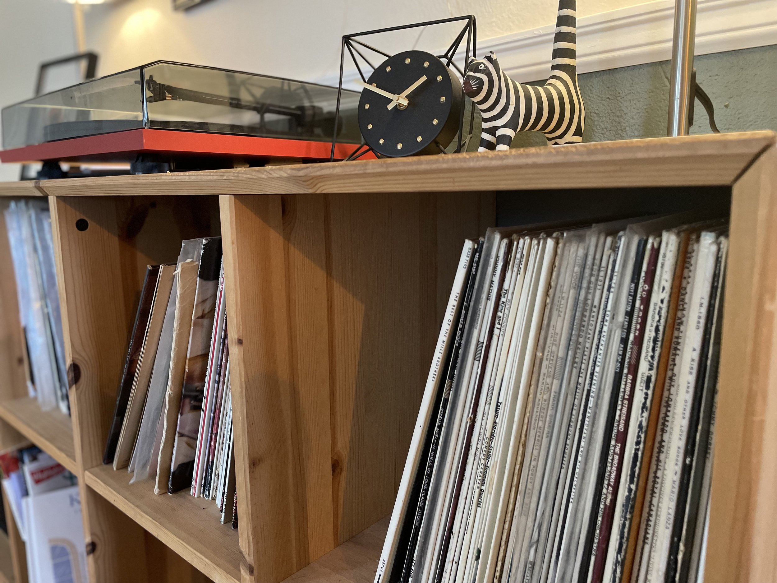 A wooden bookshelf with vinyl records, a black clock with gold hands, a zebra-shaped figurine, and a record player on top.