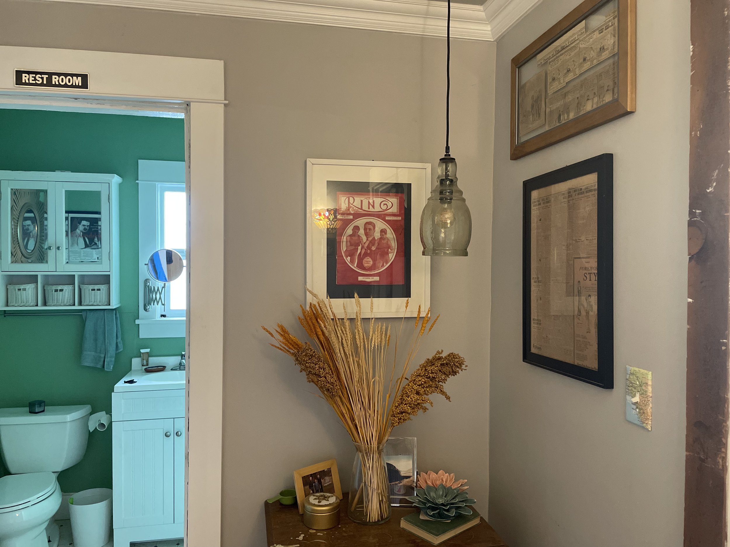 Decorative corner of a home with framed pictures on the wall, a hanging light, and a table with dried flowers, a photo frame, a succulent, and other small items.