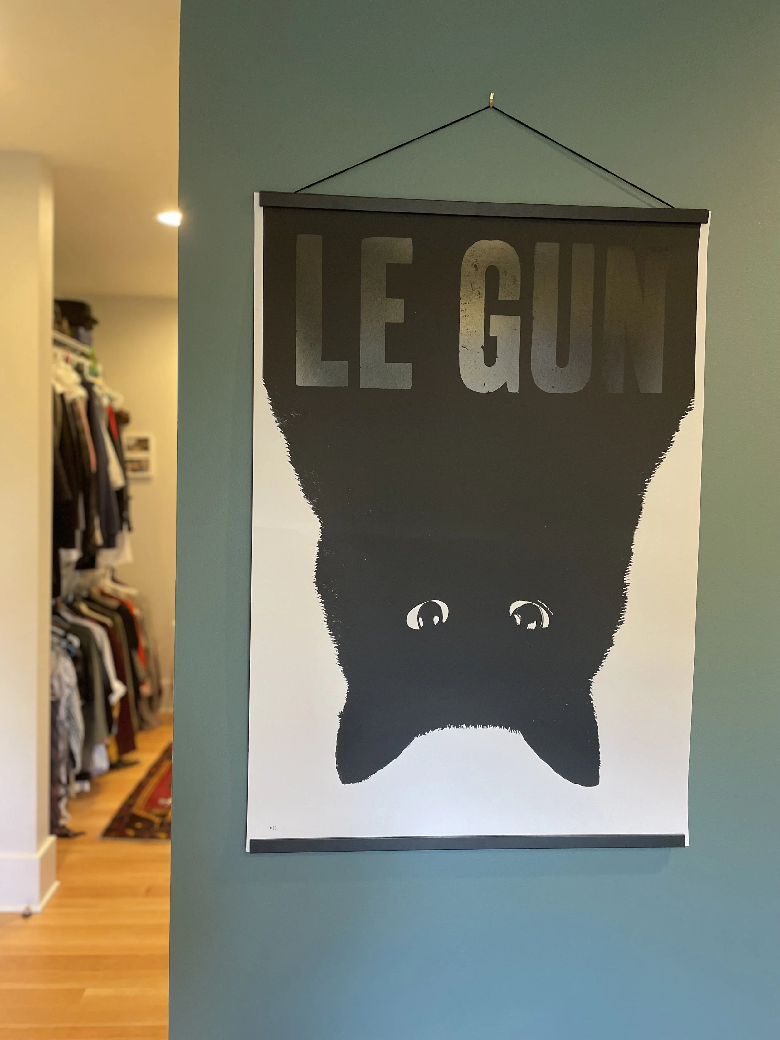 Poster of a black cat's upside-down face with the words "LE GUN" above it, hanging on a teal wall.