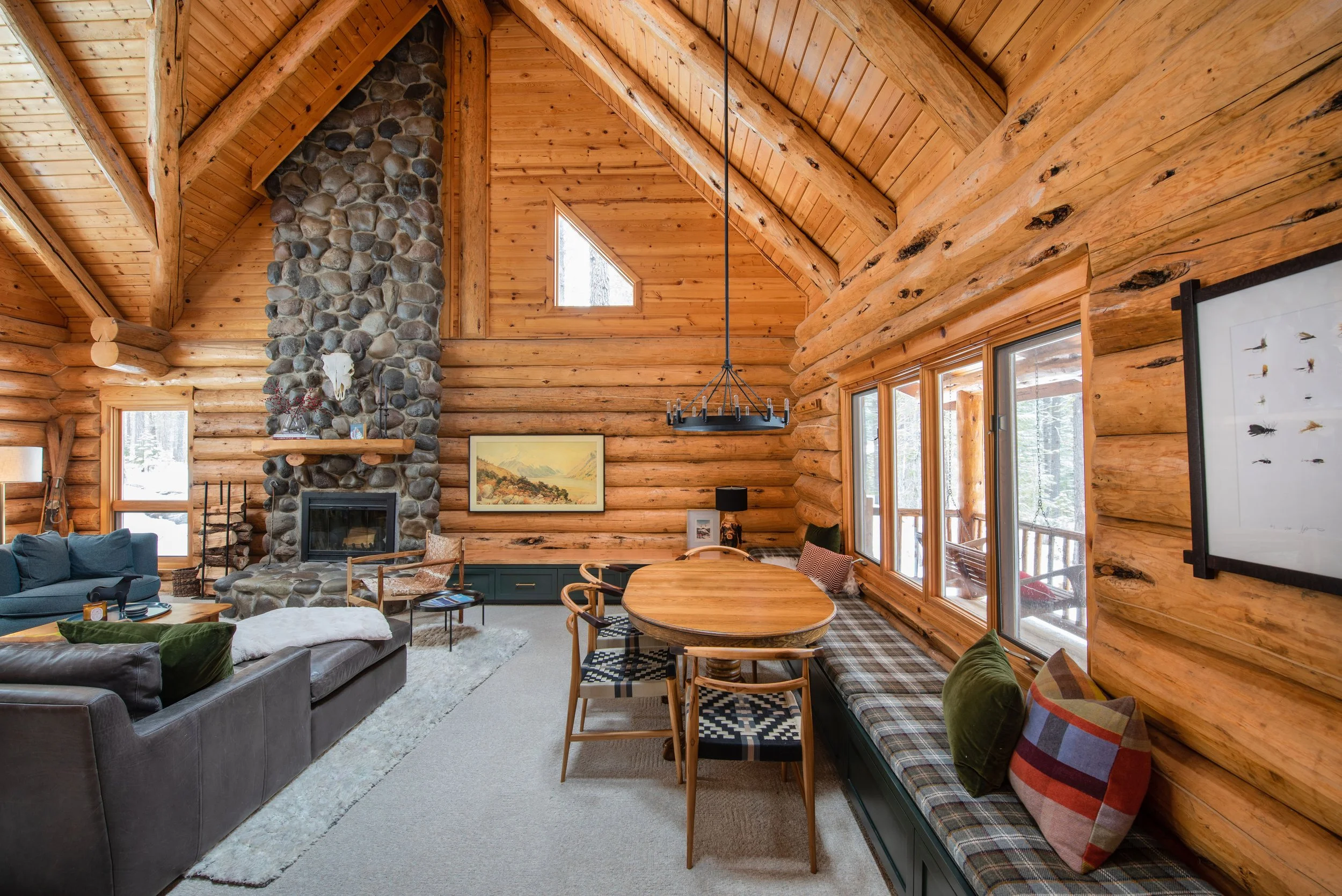 The cabin living room features modern decor, a stone fireplace, and a wrap-around bench. The media bench was added to connect two separate spaces (the game table and living room) with one side as the media wall with drawers and one side with lift-top