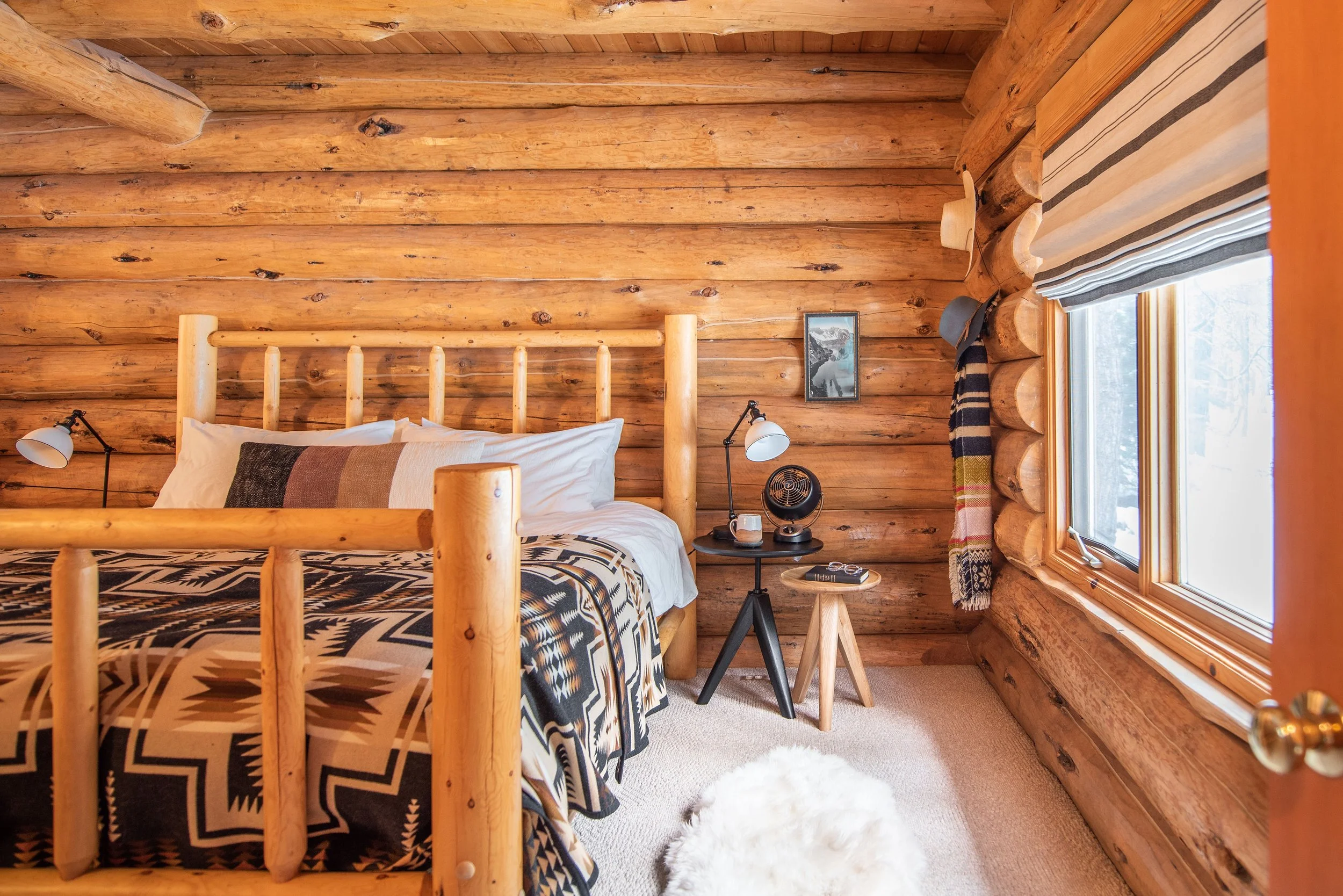 The cabin bedroom features a log bedframe with a Pendleton blanket and modern nested side tables from Rejuvenation. Vintage details like the painting of Crater Lake to the hat hooks on the wall add more personality.