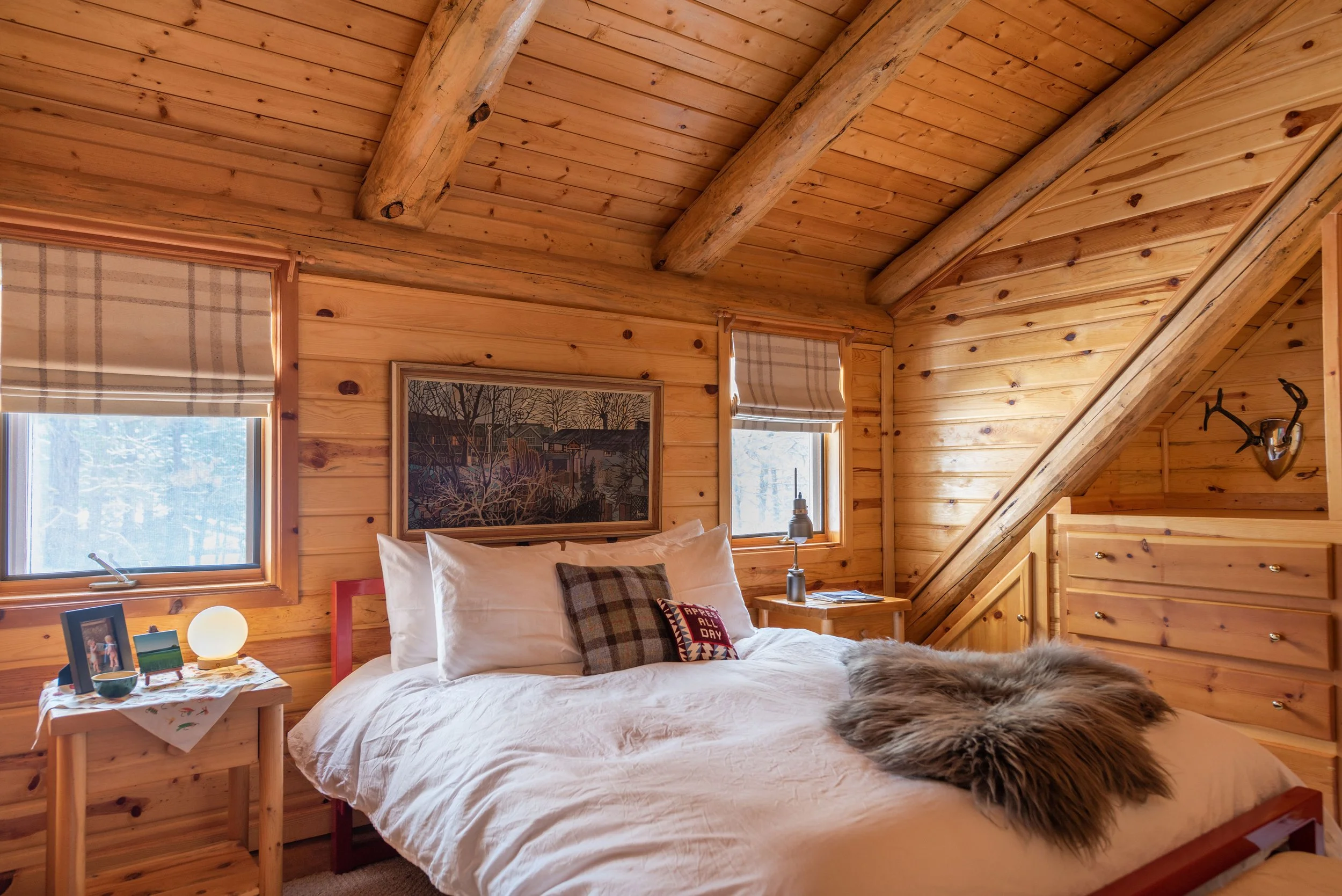 This cabin bedroom with log walls and crisp white bedding features a painting passed down from the owner's late father and beautiful curtains from The Shade Store.