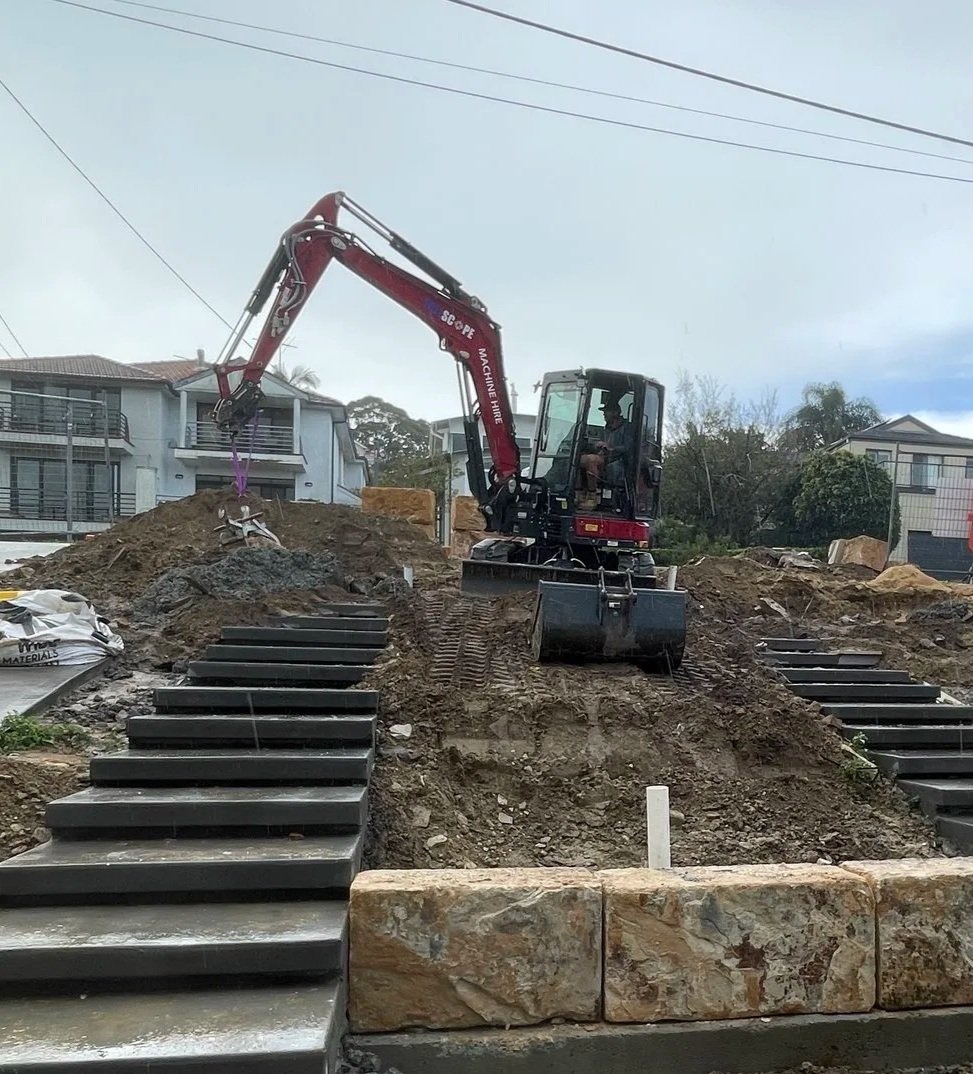 A construction site with a small excavator on a dirt hill, surrounded by residential buildings and power lines in the background.