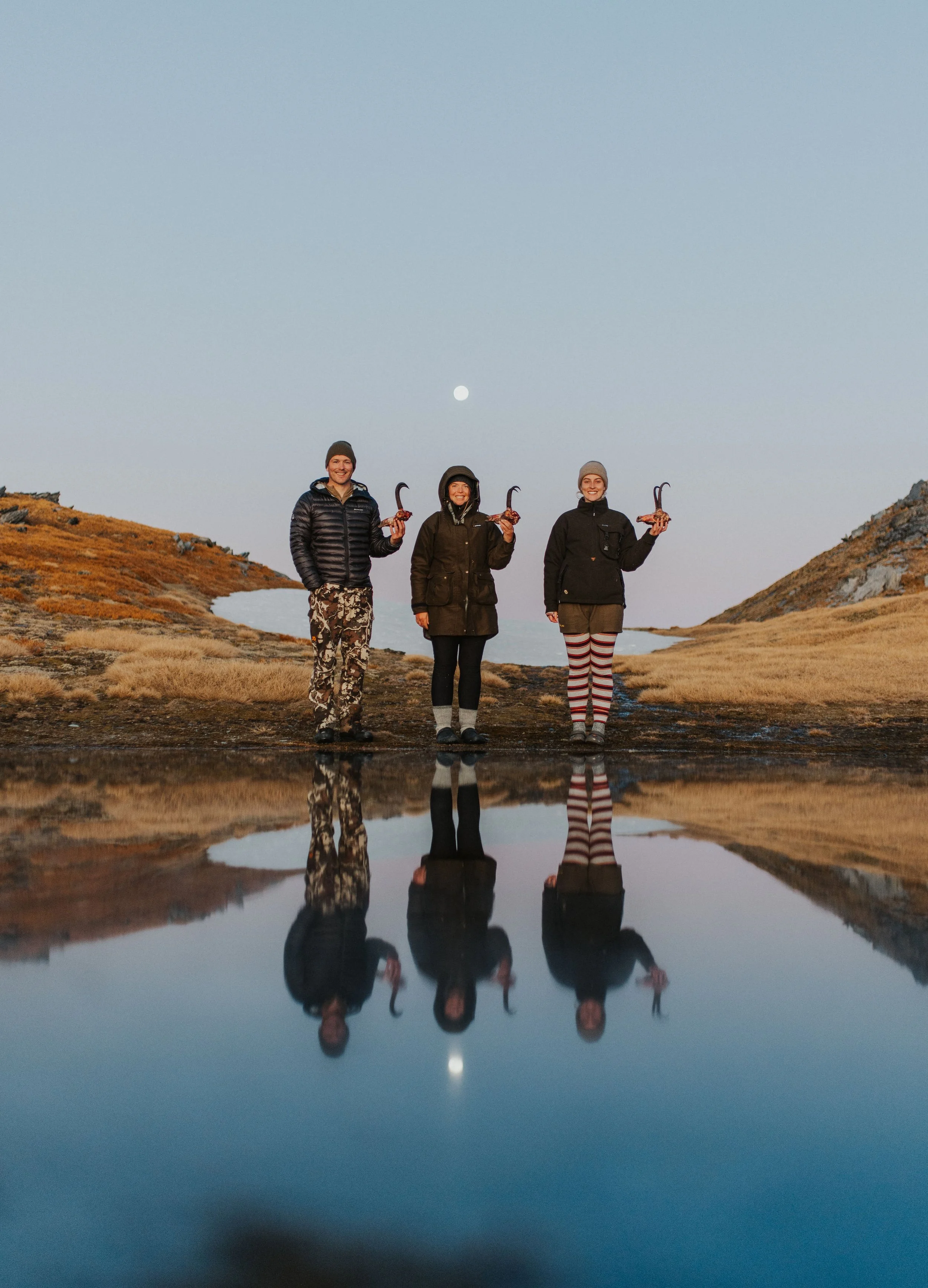 Three people standing on rocky terrain near a body of water at dusk, holding chamois trophies with a full moon in the sky reflected in the water.