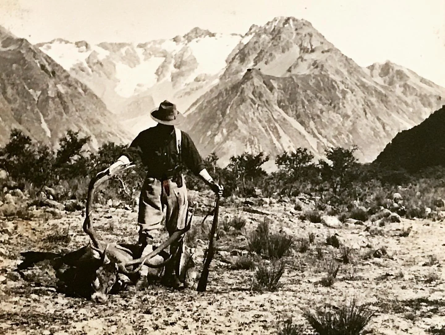 A person in outdoor clothing and a wide-brimmed hat stands with mountains in the background, holding a .red stag's antlers