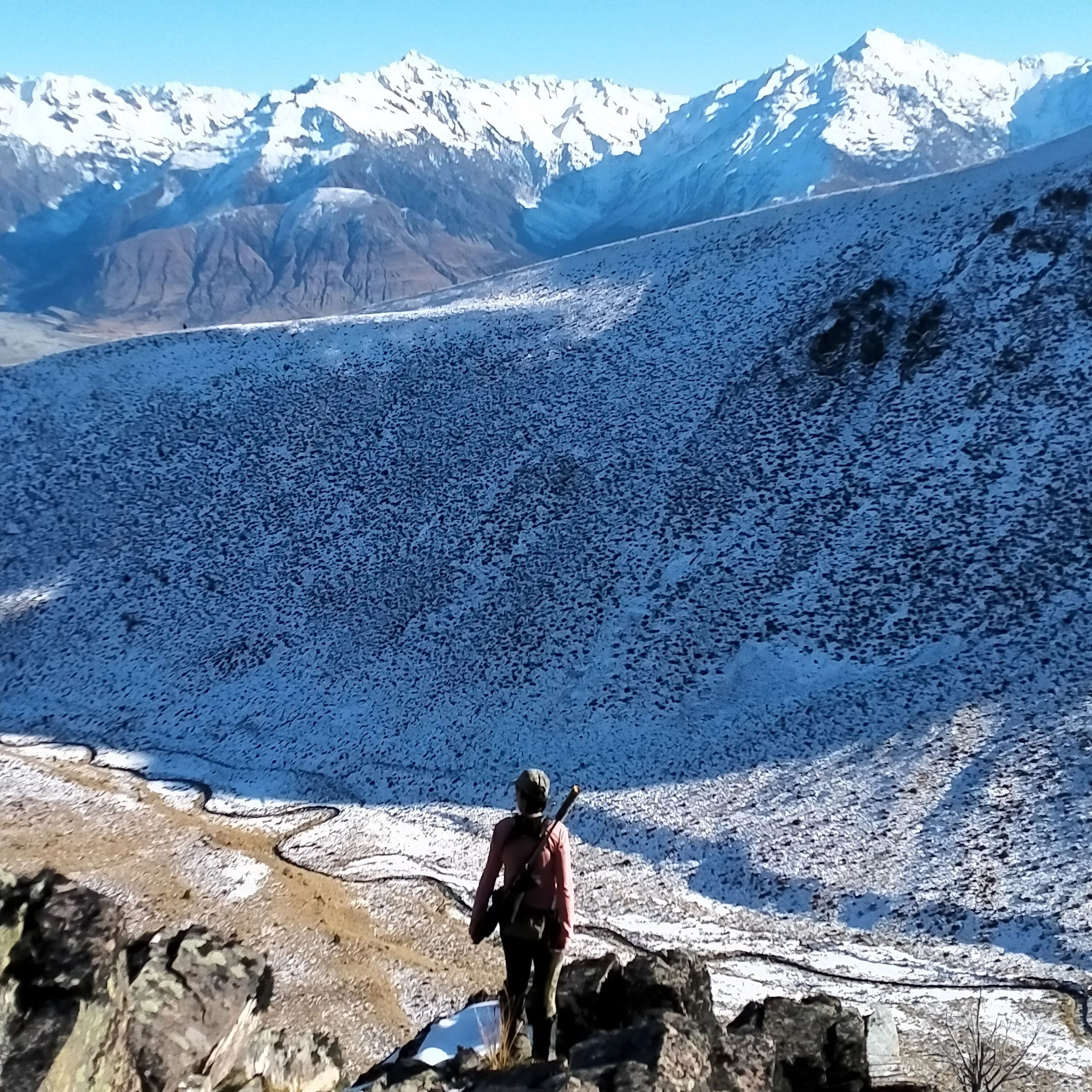 Person in hunting camoflague standing on rocky ground near a large body of water with snow-capped mountains in the background.