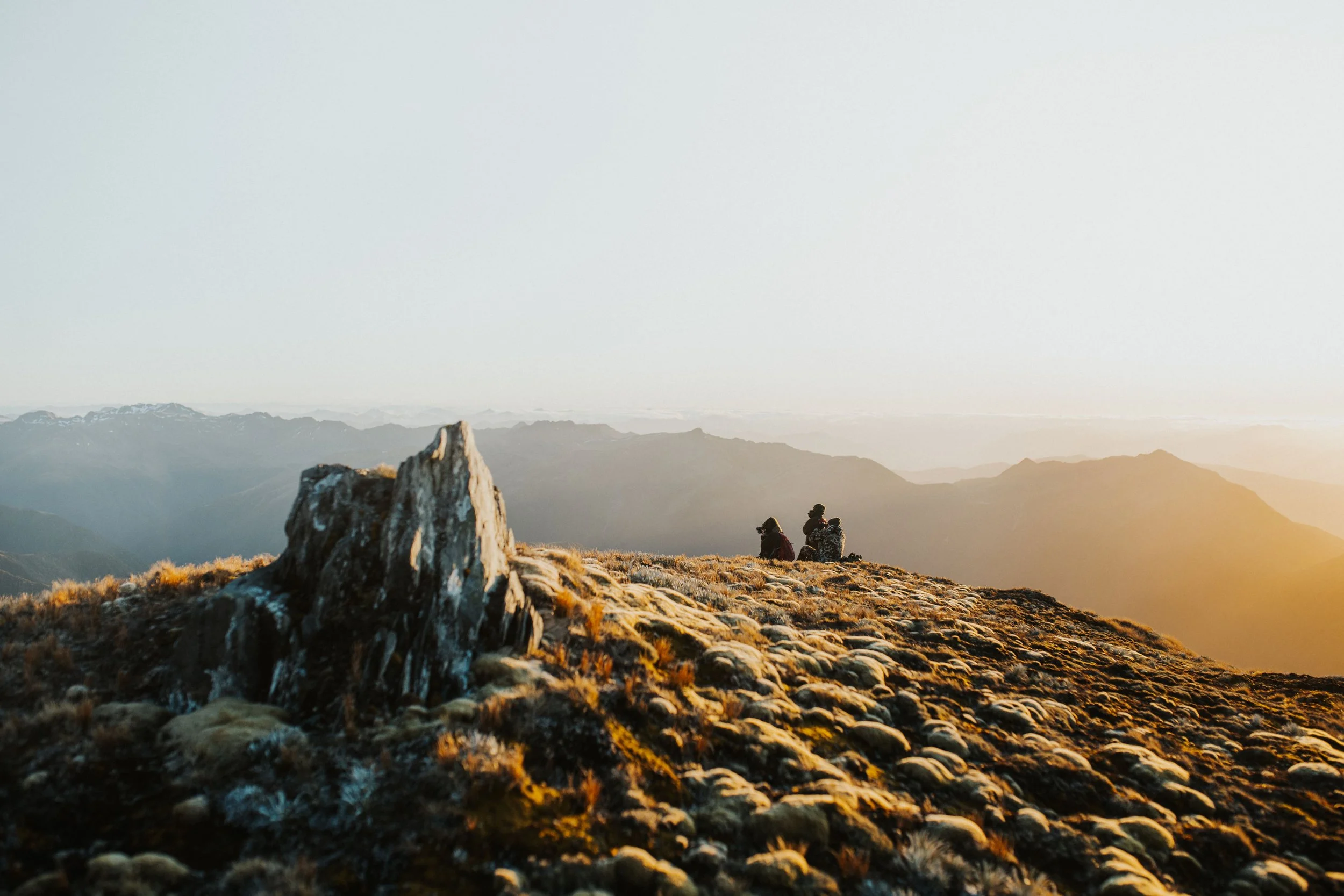 Two hikers sitting and enjoying the view of a mountain landscape at sunset.