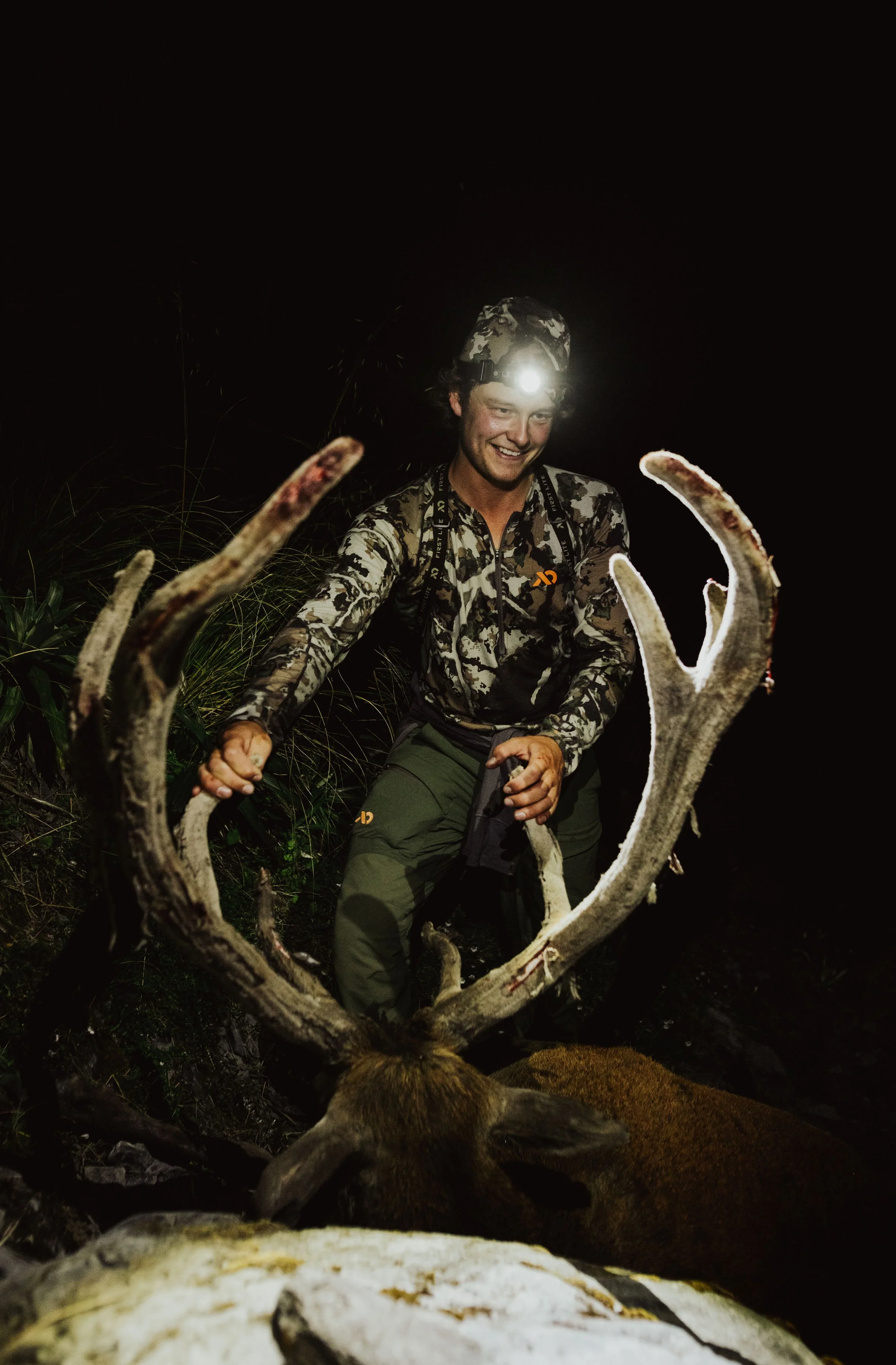 A person wearing camouflage clothing, and a headlamp,  is smiling behind a set of large antlers at night.