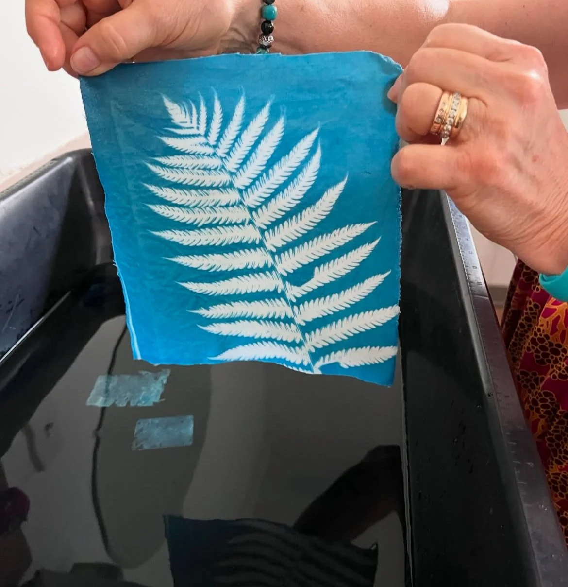 Person holding a piece of blue paper with a white fern leaf print, with a sink and water in the background.